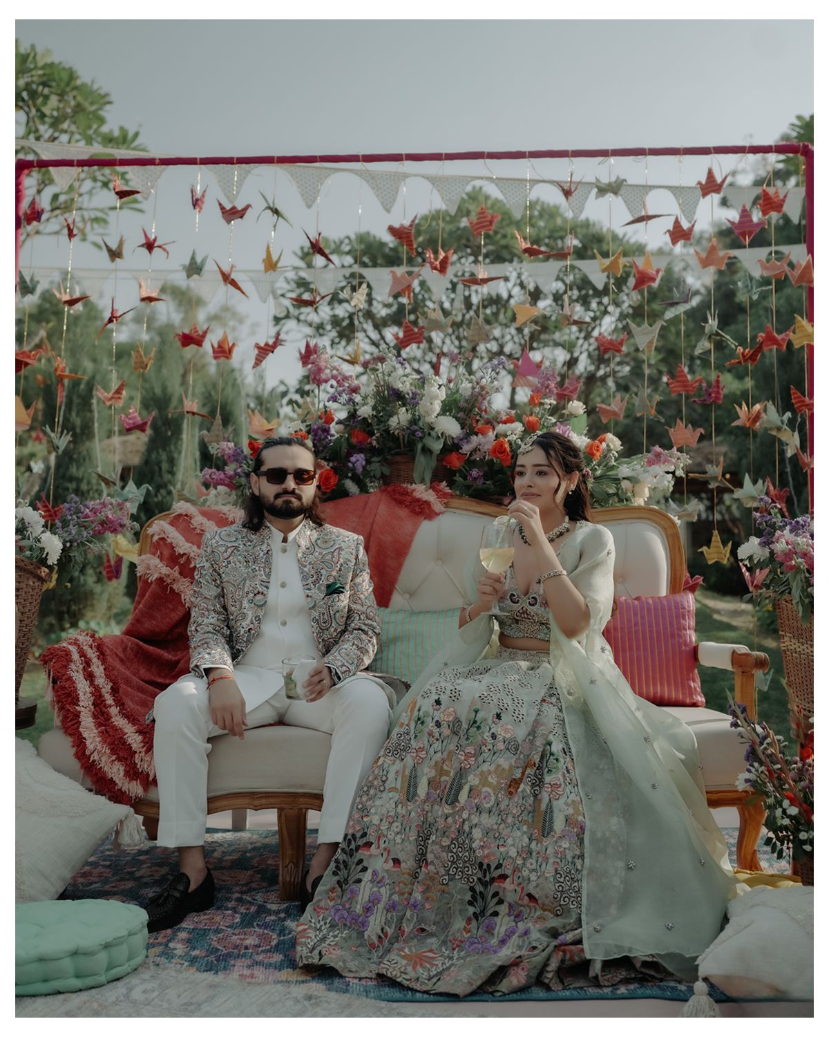 A couple sits together on an elegant loveseat surrounded by a stunning floral wedding display with hanging decorations.