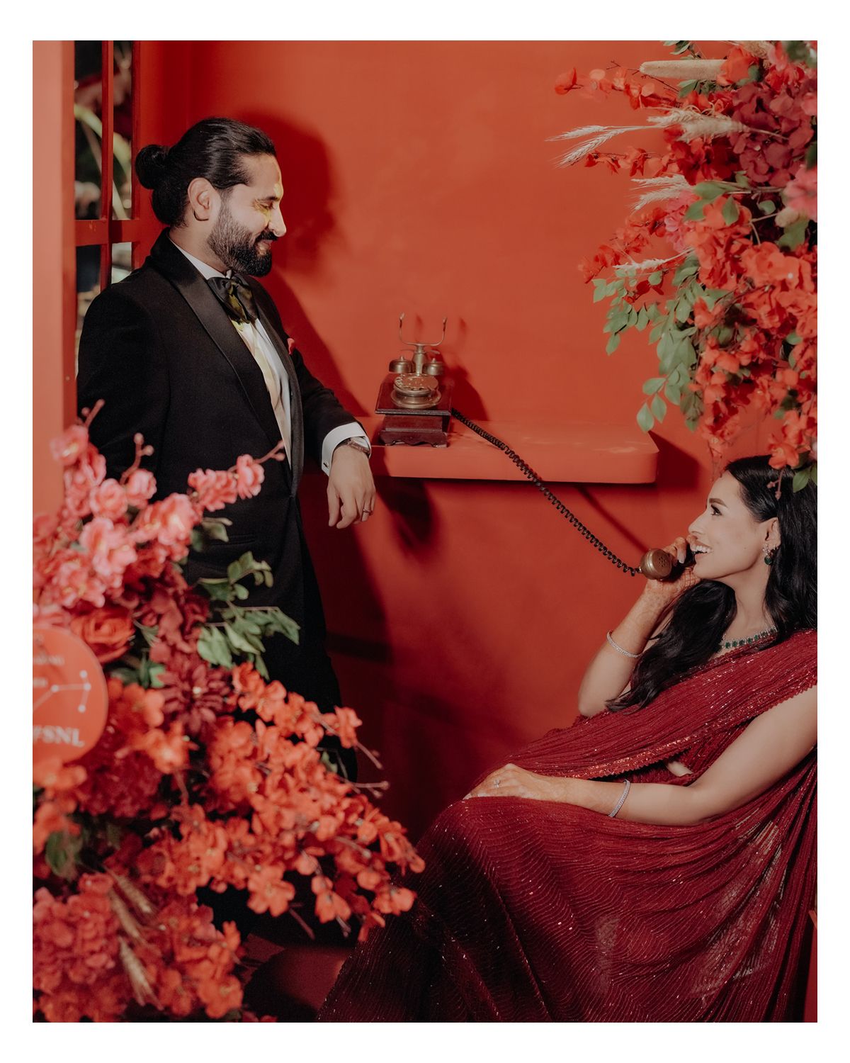 Stylish couple poses in vintage-inspired scene with red wall and pink floral decorations against dramatic lighting.