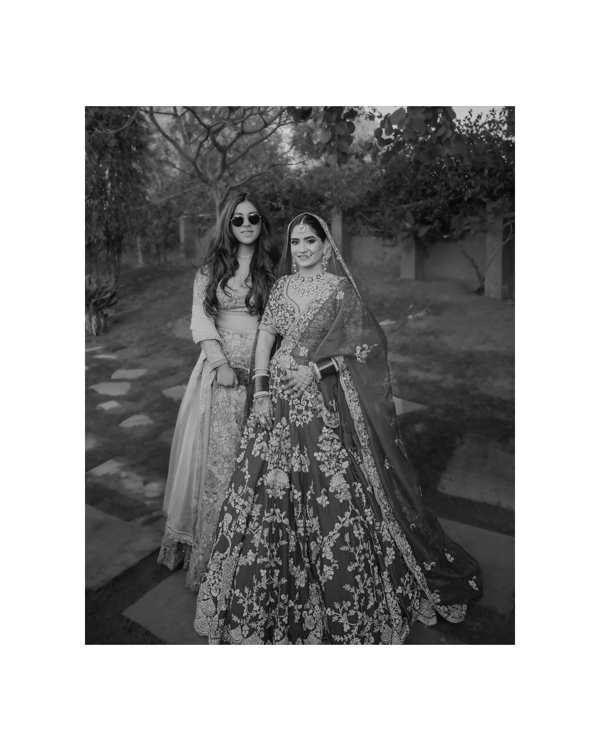 Two women in elegant Indian wedding attire pose together in a black and white outdoor portrait.
