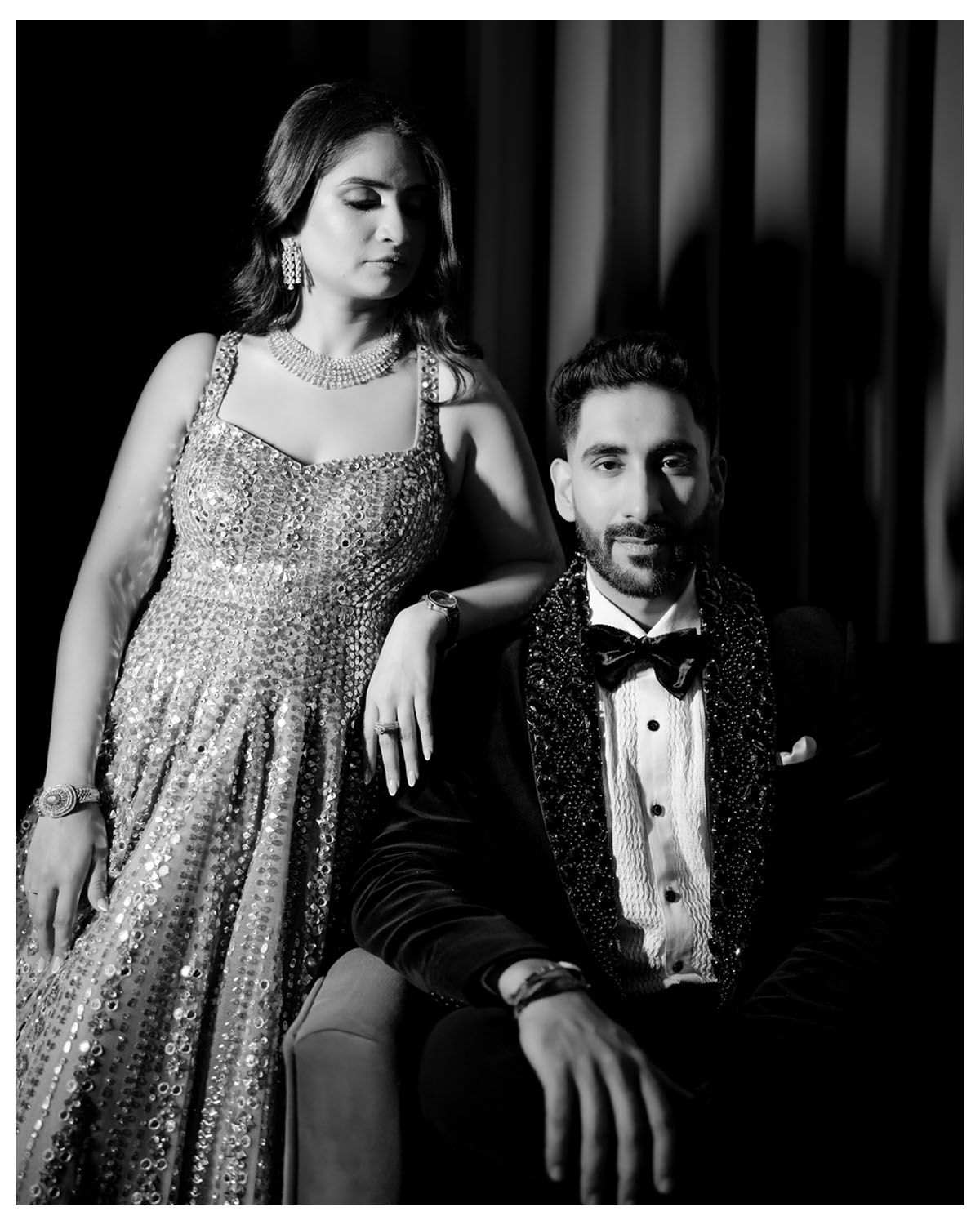 Black and white portrait of couple in formal wear seated on couch with dramatic lighting.