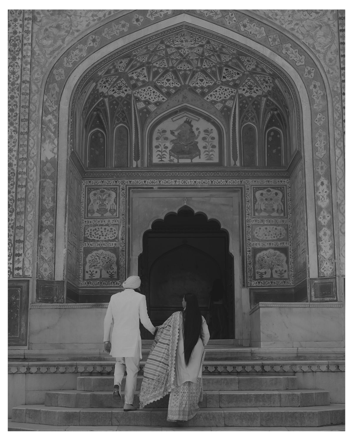 A couple walking up marble steps towards an elaborately decorated Mughal-style archway entrance.