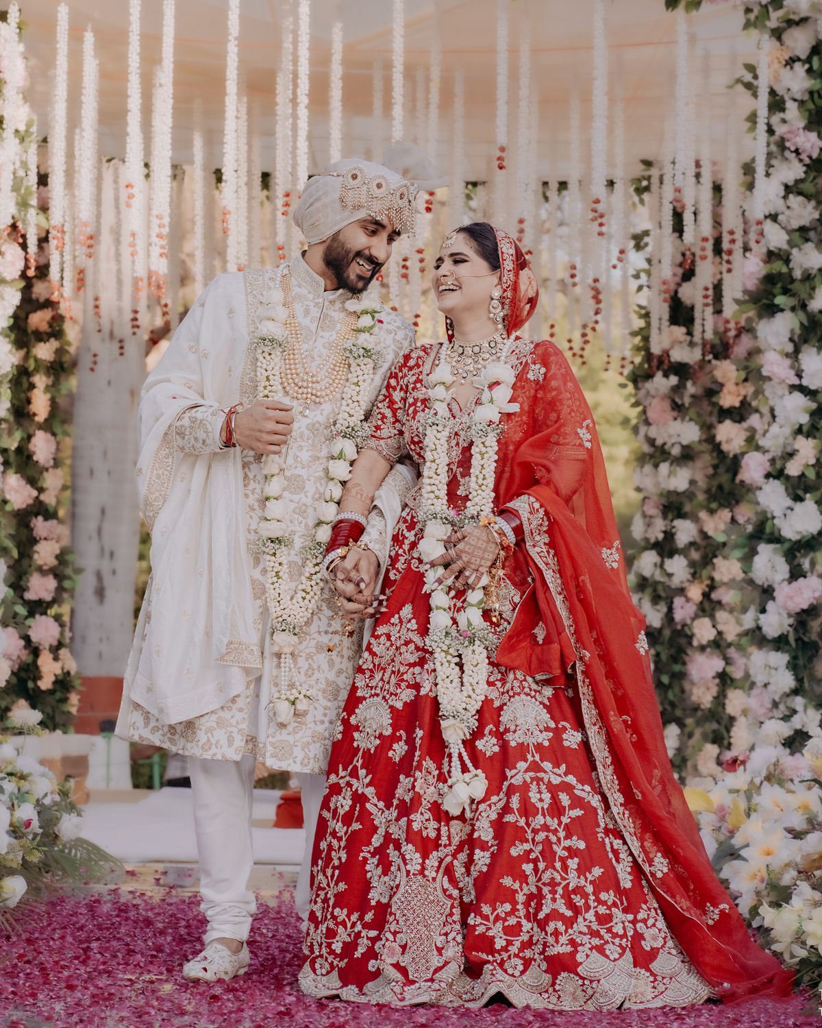 Couple poses under floral mandap with hanging white decorations in traditional wedding attire.