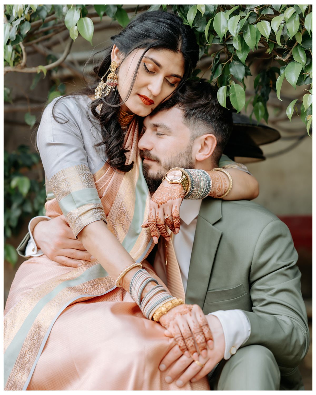 Romantic couple in traditional South Asian attire sharing an intimate moment against a leafy backdrop.