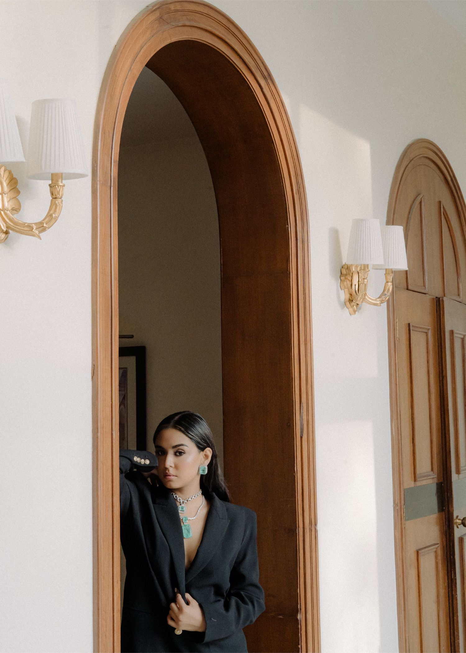 A person in a dark blazer poses against a beige wall with arched doorways in an elegant interior setting.