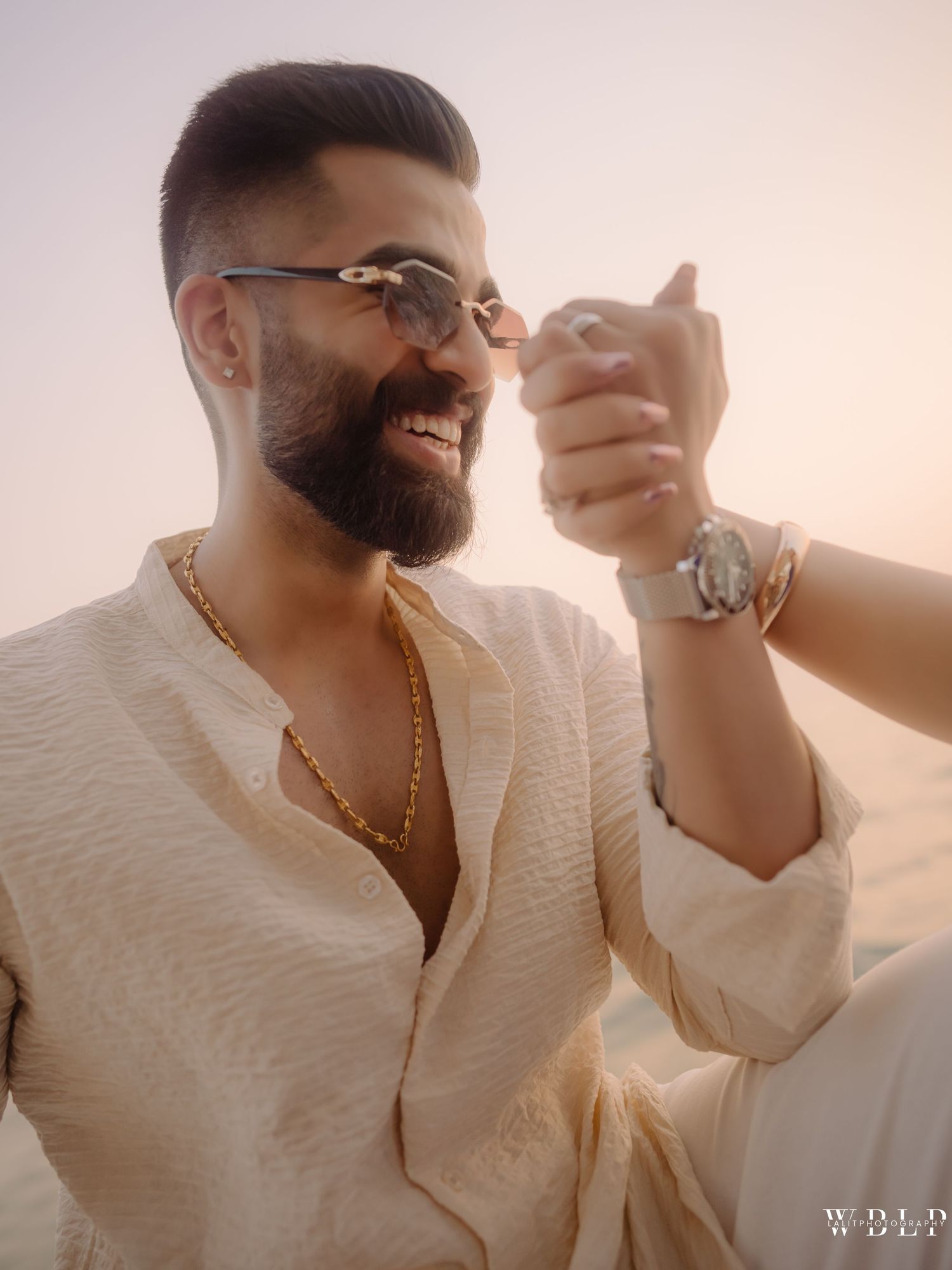 Smiling bearded man in cream kurta with gold chain and watch, holding sunglasses outdoors at sunset.