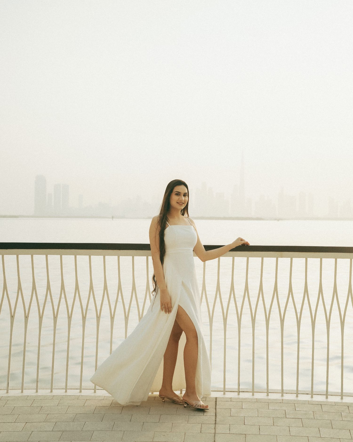 Woman in white slit gown posing on waterfront promenade with city skyline backdrop.