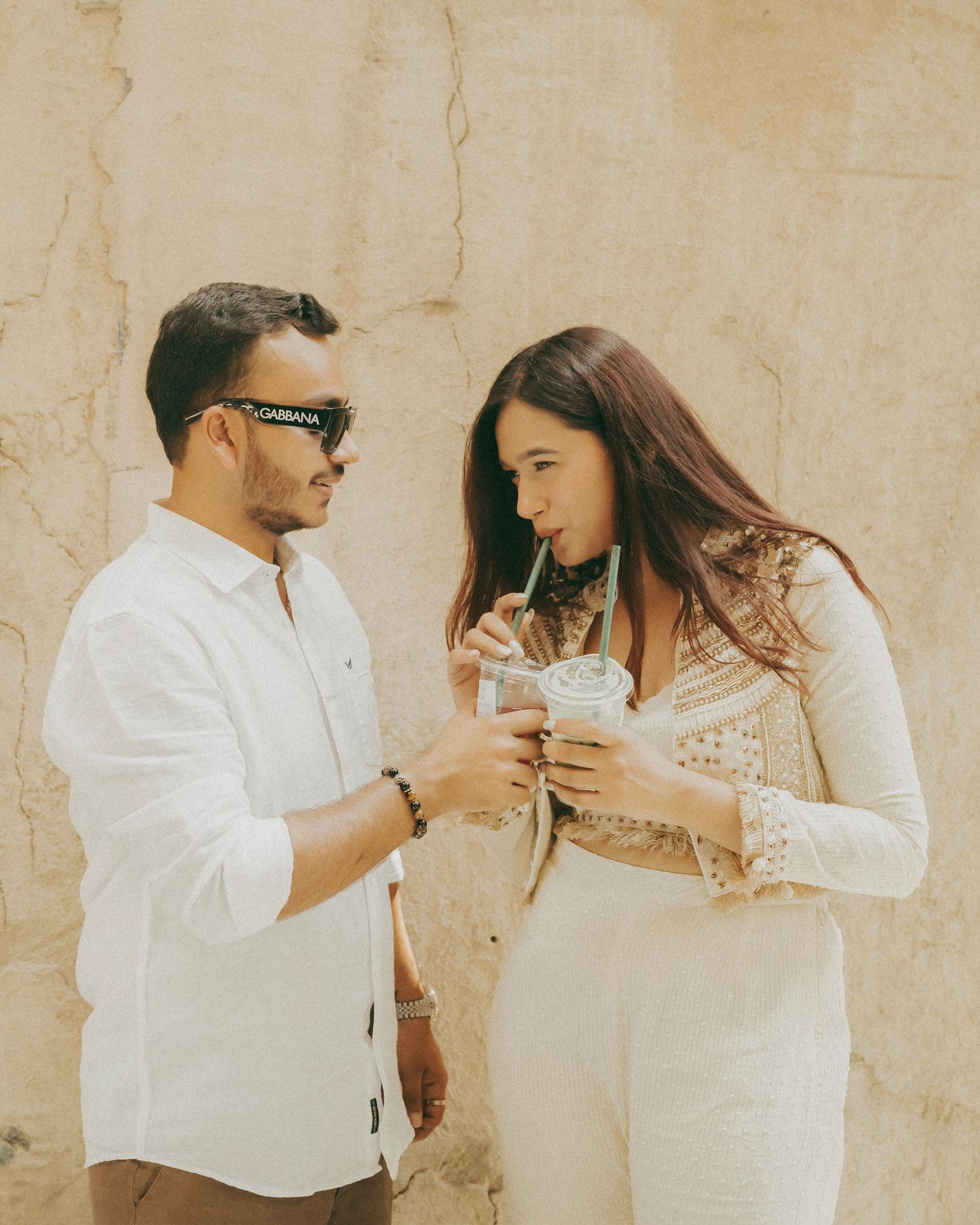 Couple in white outfits sharing drinks against a beige stone wall outdoors.