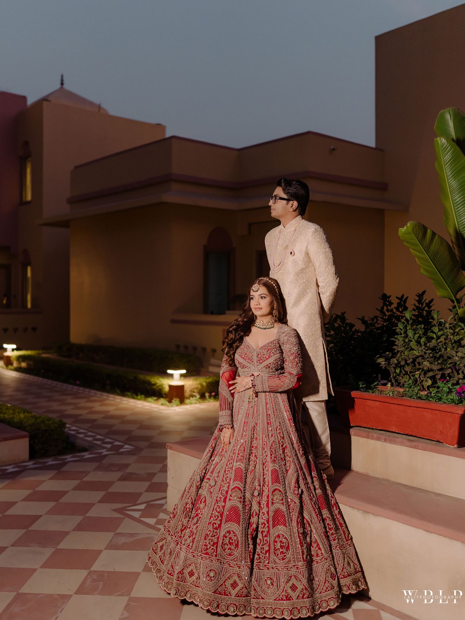 Indian bride in red lehenga and groom in white sherwani pose elegantly outside a venue at dusk.