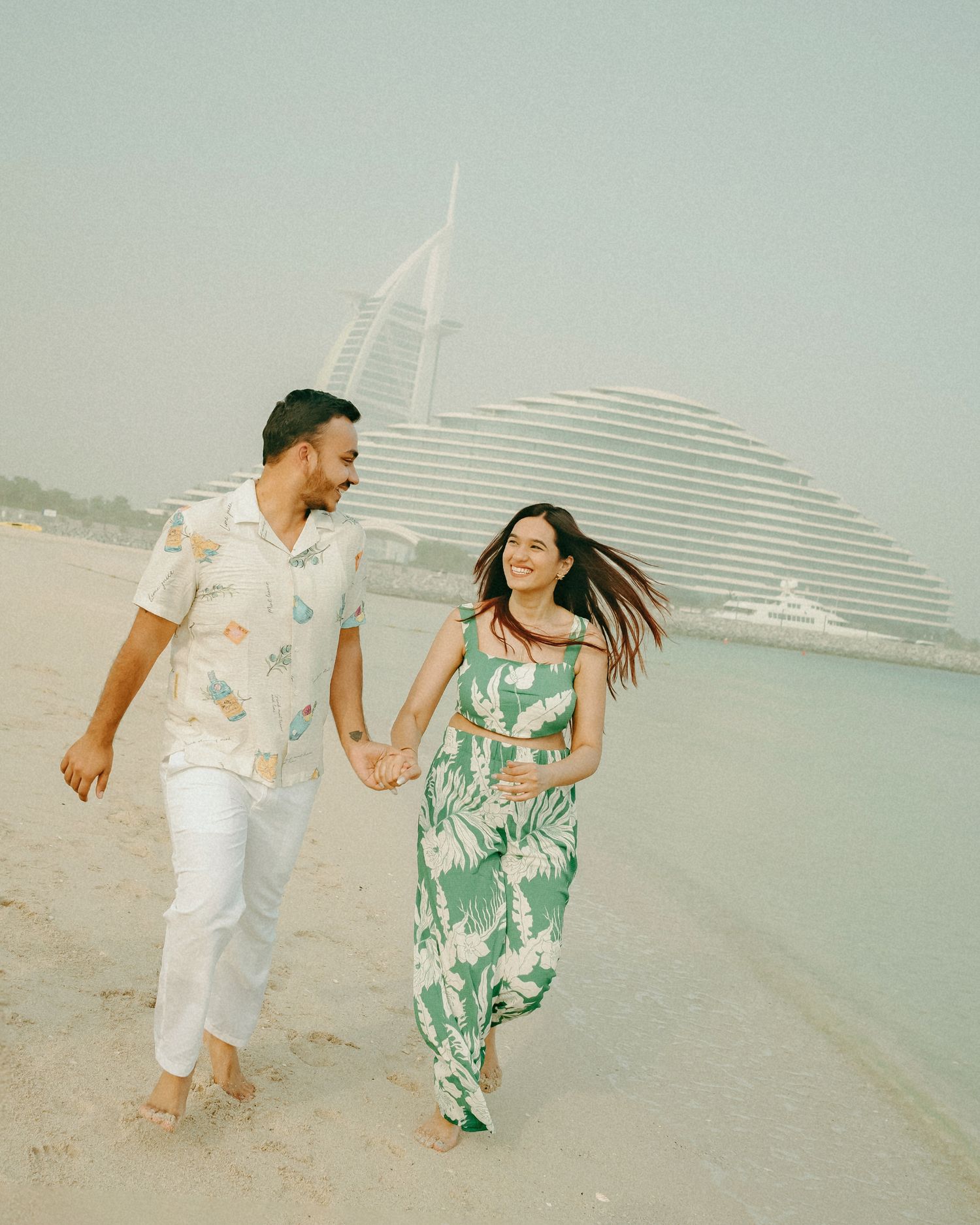 Couple holding hands walking on Dubai beach with iconic Burj Al Arab hotel in background.