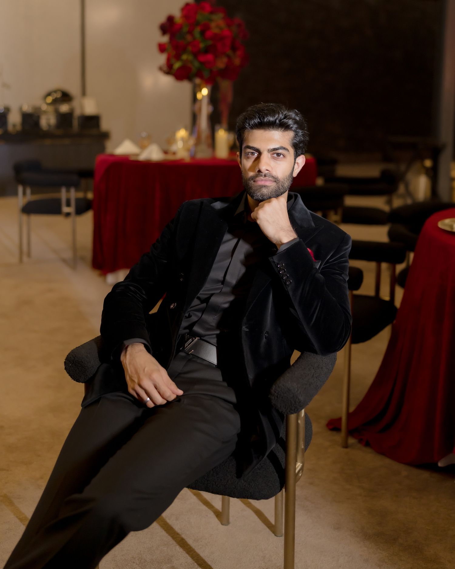 A well-dressed man in a black velvet suit sits confidently on a chair in an elegant red and black decorated venue.