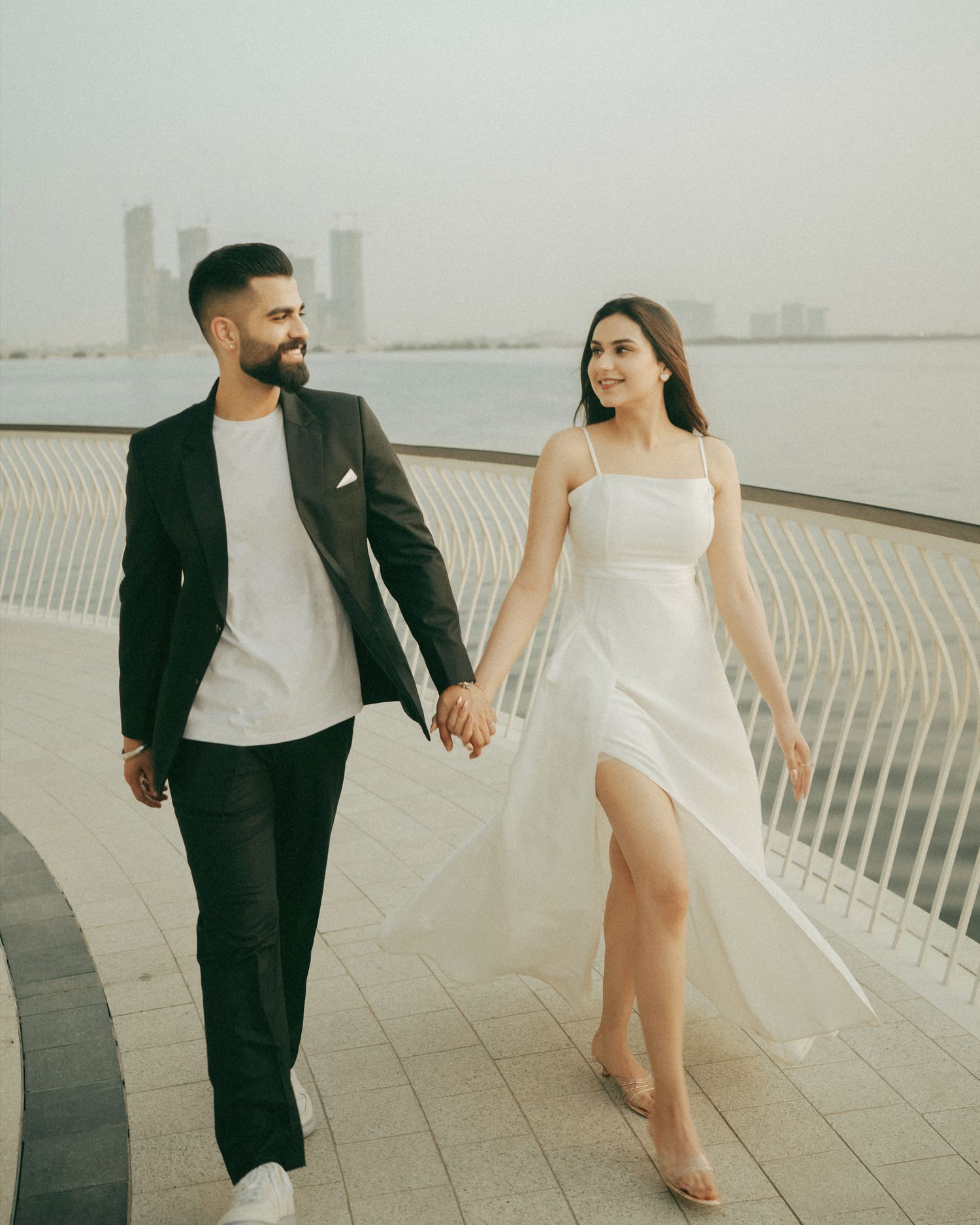 Couple holding hands walking along waterfront promenade, man in black suit, woman in white dress.
