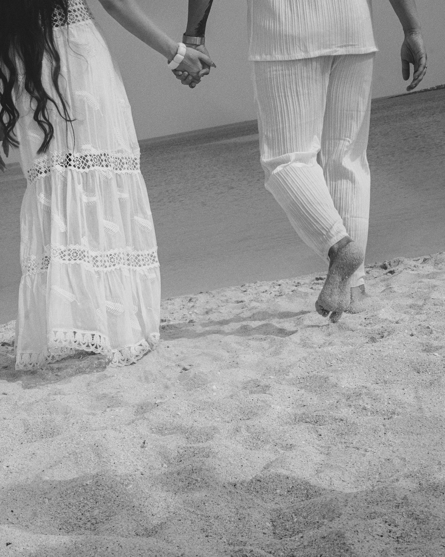 Black and white photo of a couple holding hands on sandy beach, woman in white lace dress, man in white linen pants.