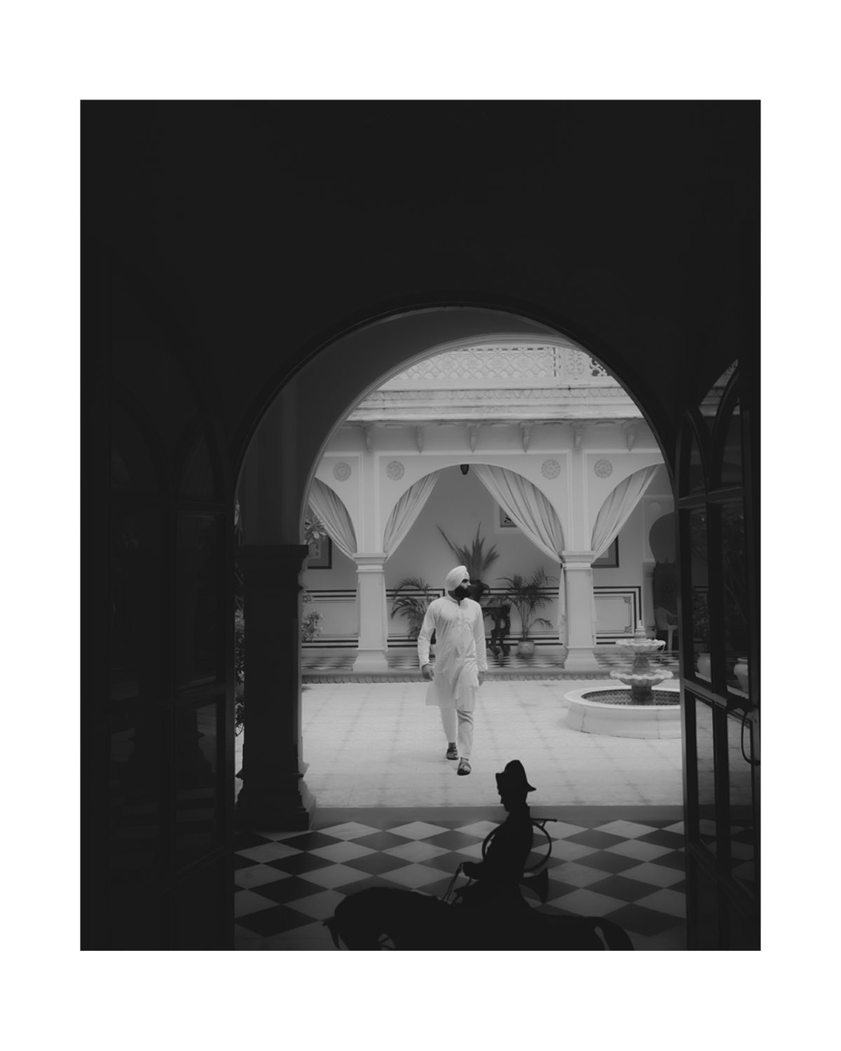 Black and white photo of arched corridor with checkered floor and distant figure.