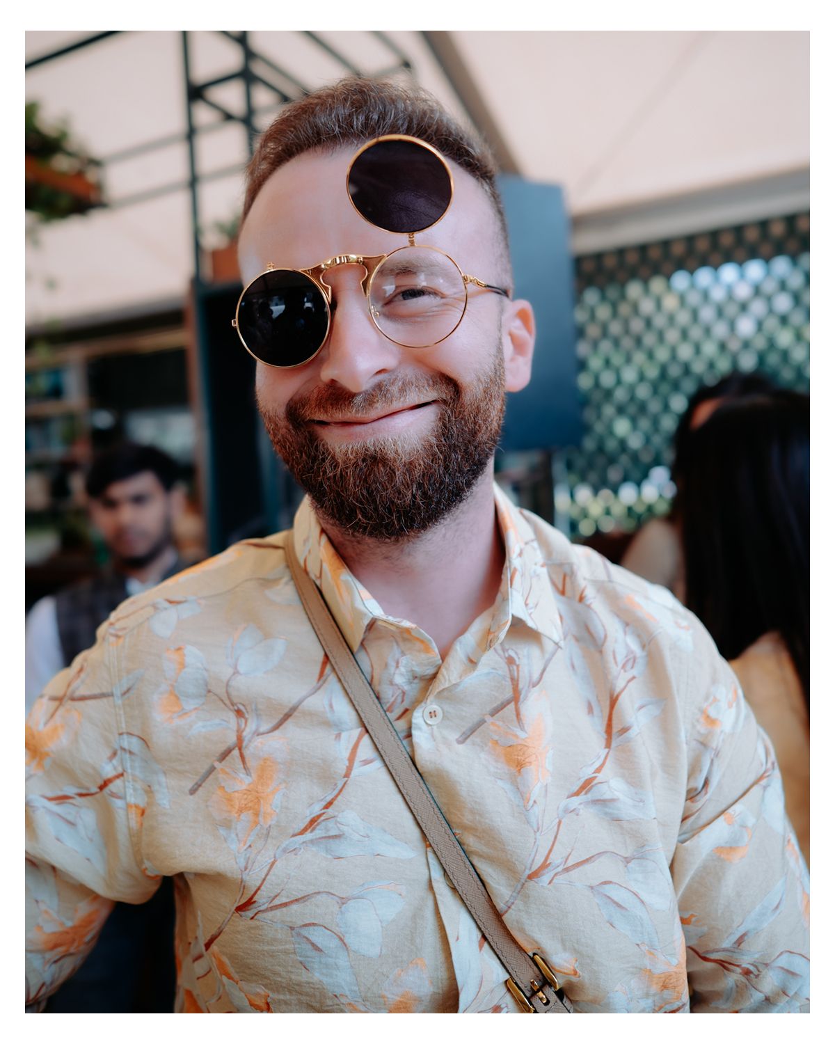A smiling person wearing sunglasses, glasses, and a floral print short sleeve shirt outdoors.