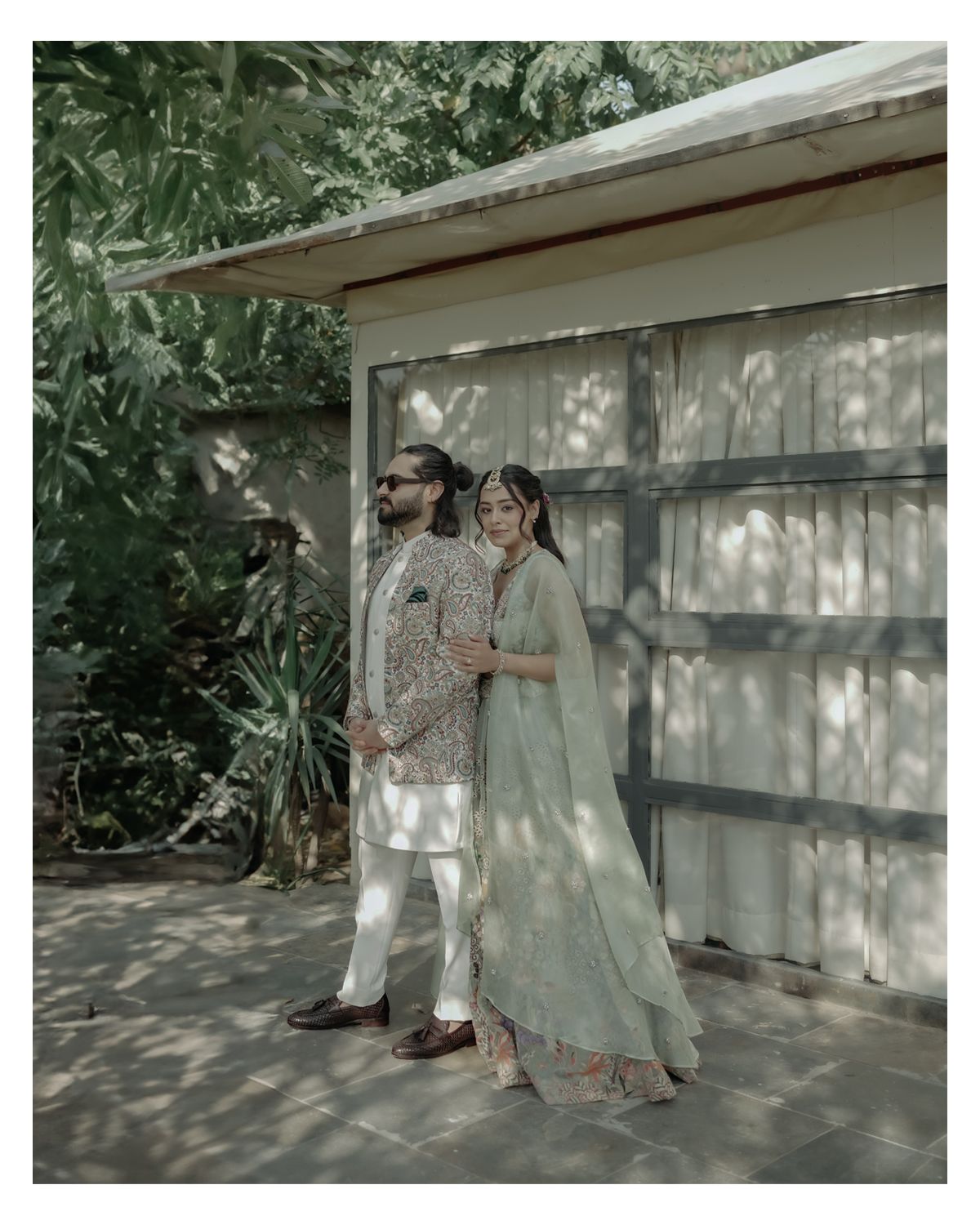 A couple in traditional South Asian attire poses together against a white exterior wall in dappled sunlight.