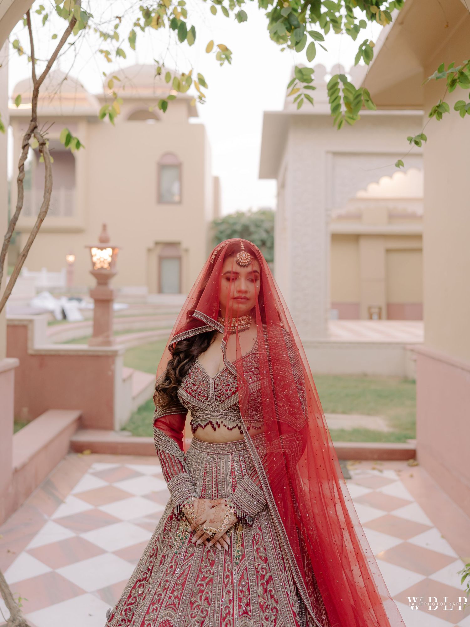 Bride in red lehenga with dupatta poses at a palace courtyard in traditional Indian bridal attire.