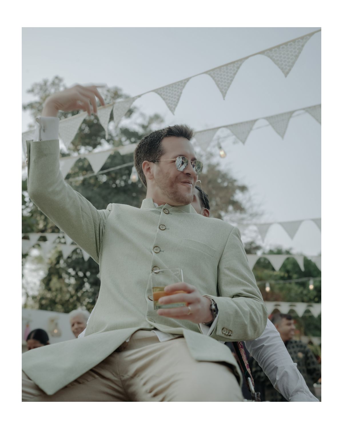 A person in a beige suit jacket raises a glass under white bunting decorations at an outdoor celebration.
