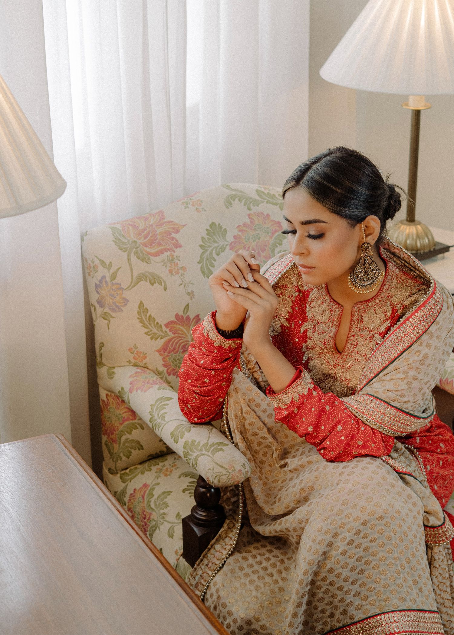 A person in traditional South Asian attire with ornate jewelry sits contemplatively on a floral patterned couch near table lamps.