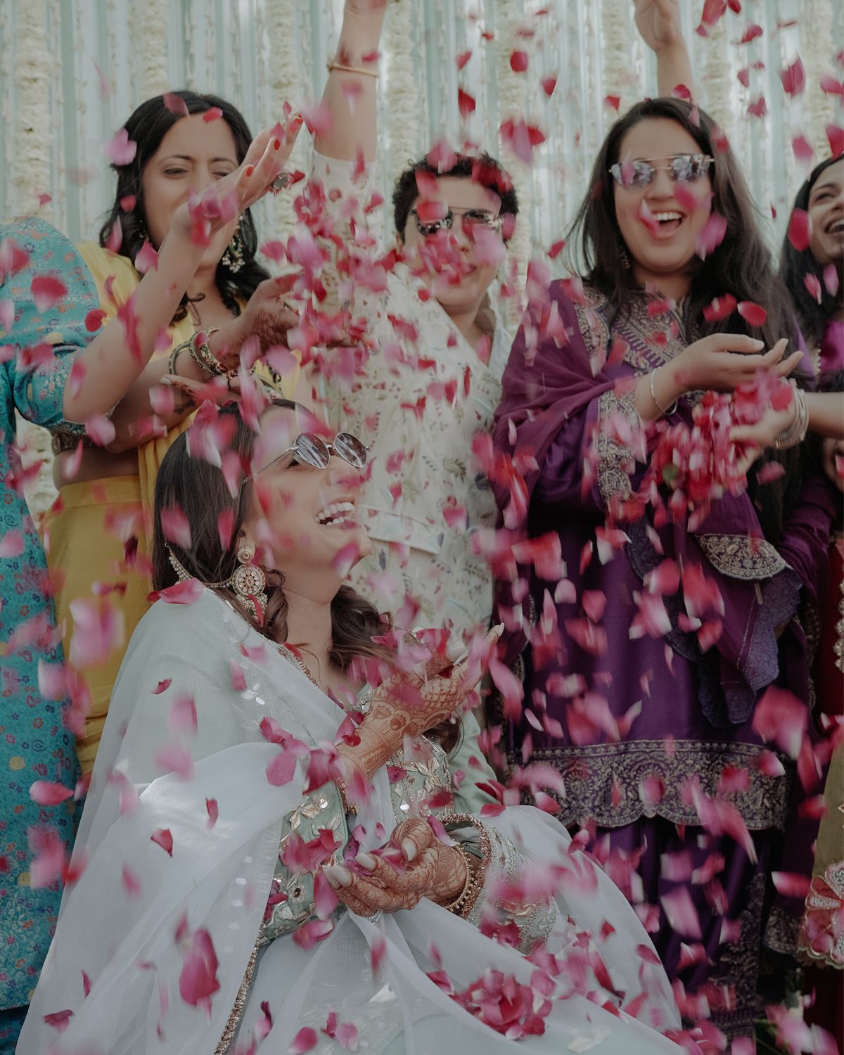 Candid moment of bride and friends laughing amidst shower of colorful rose petals.