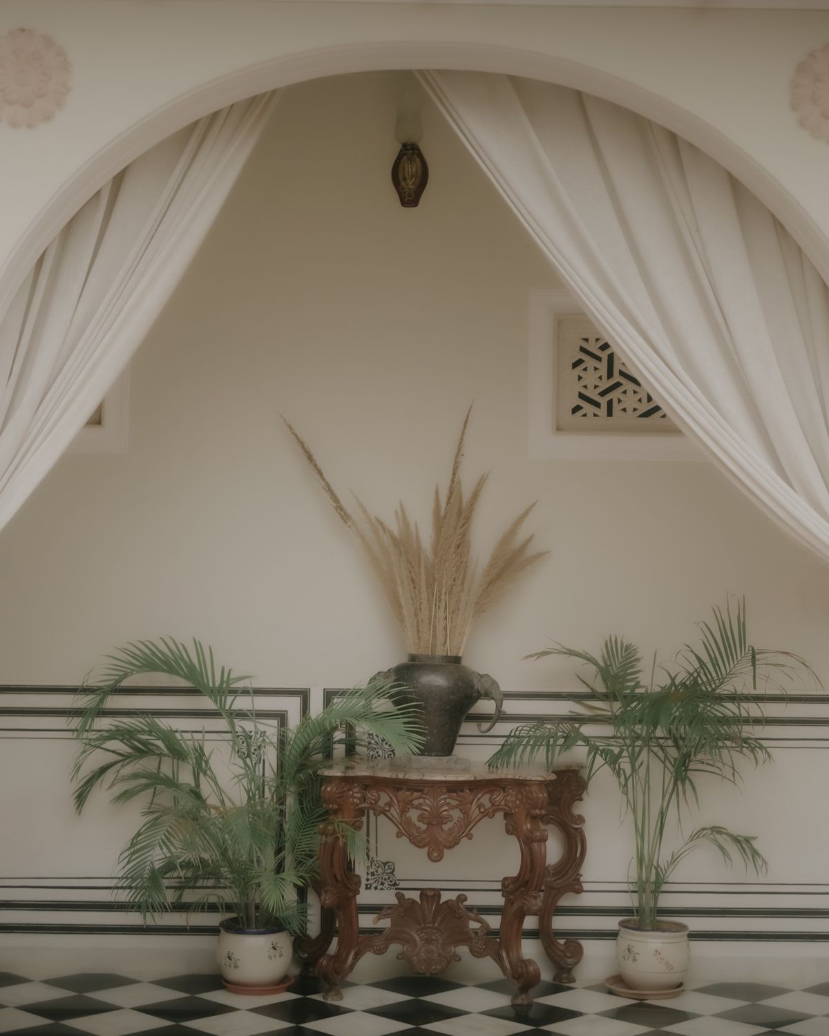 Elegant interior alcove with white drapes, potted palms, and ornate wooden console table.