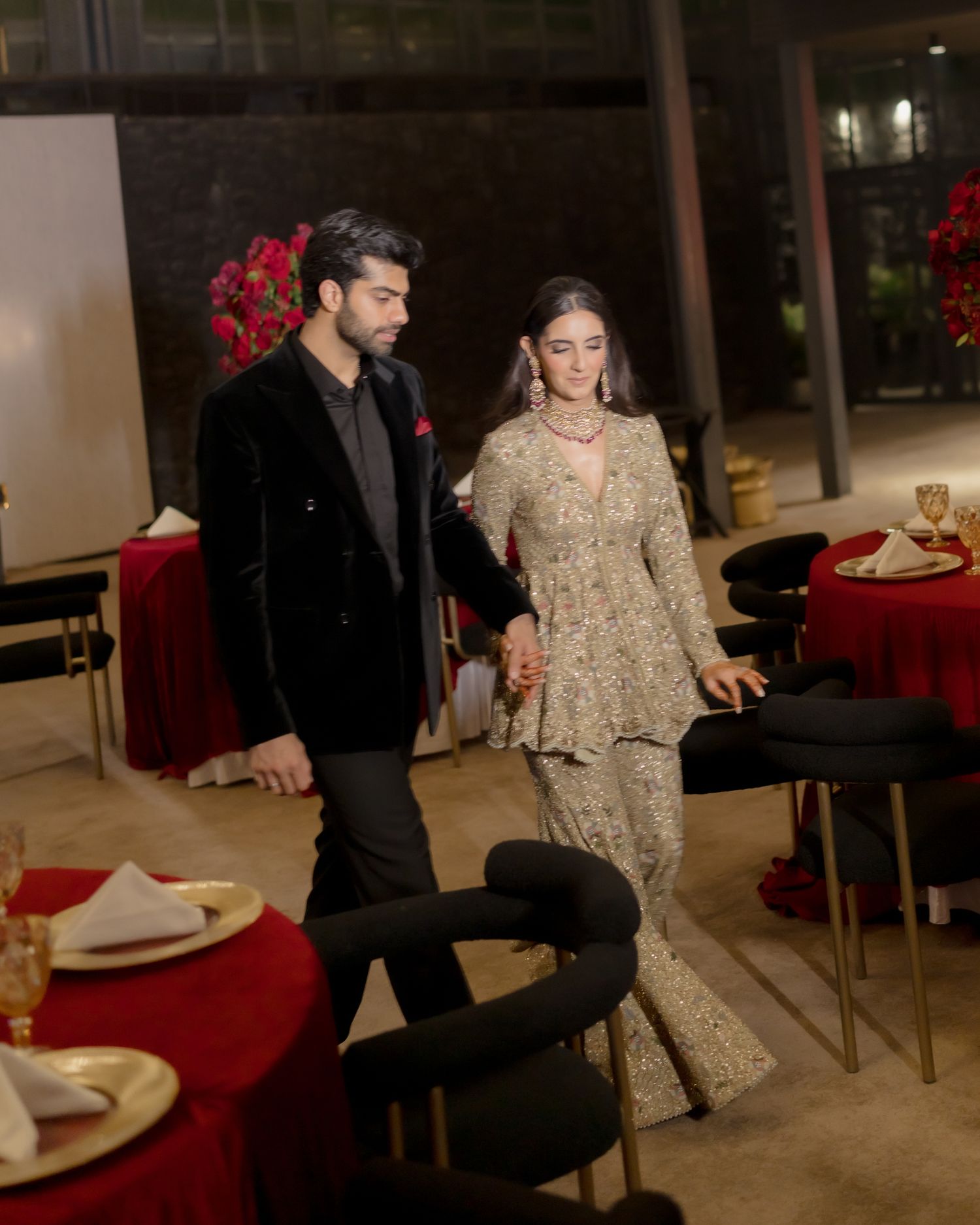 Elegant couple in formal South Asian attire holding hands at a decorated event venue with red floral arrangements.