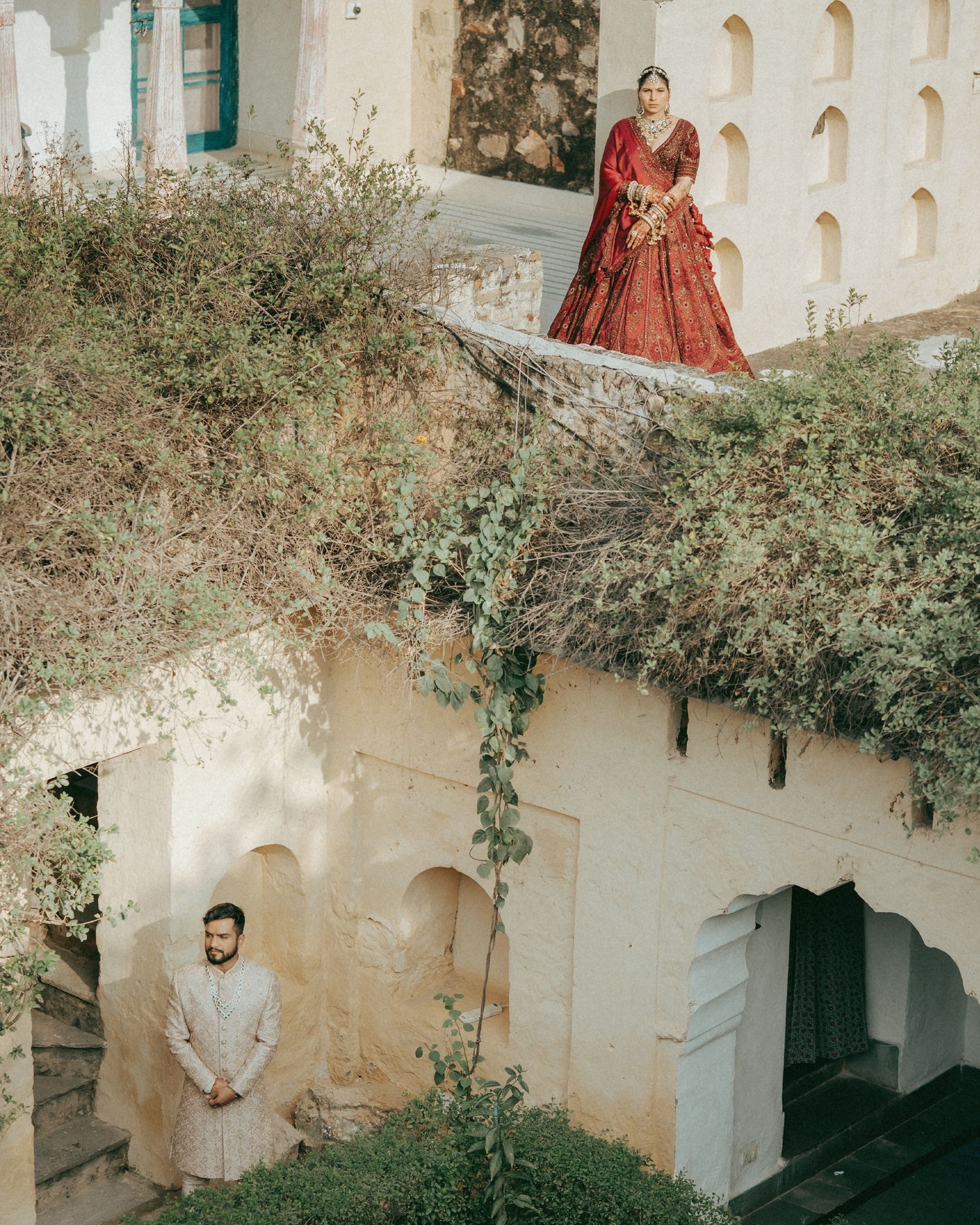Indian bride in red lehenga stands on terrace above groom in white sherwani at a heritage palace wedding.