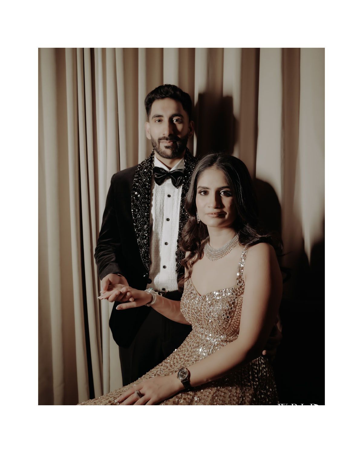Formal couple portrait with man in tuxedo and woman in sparkly gown against curtain backdrop.