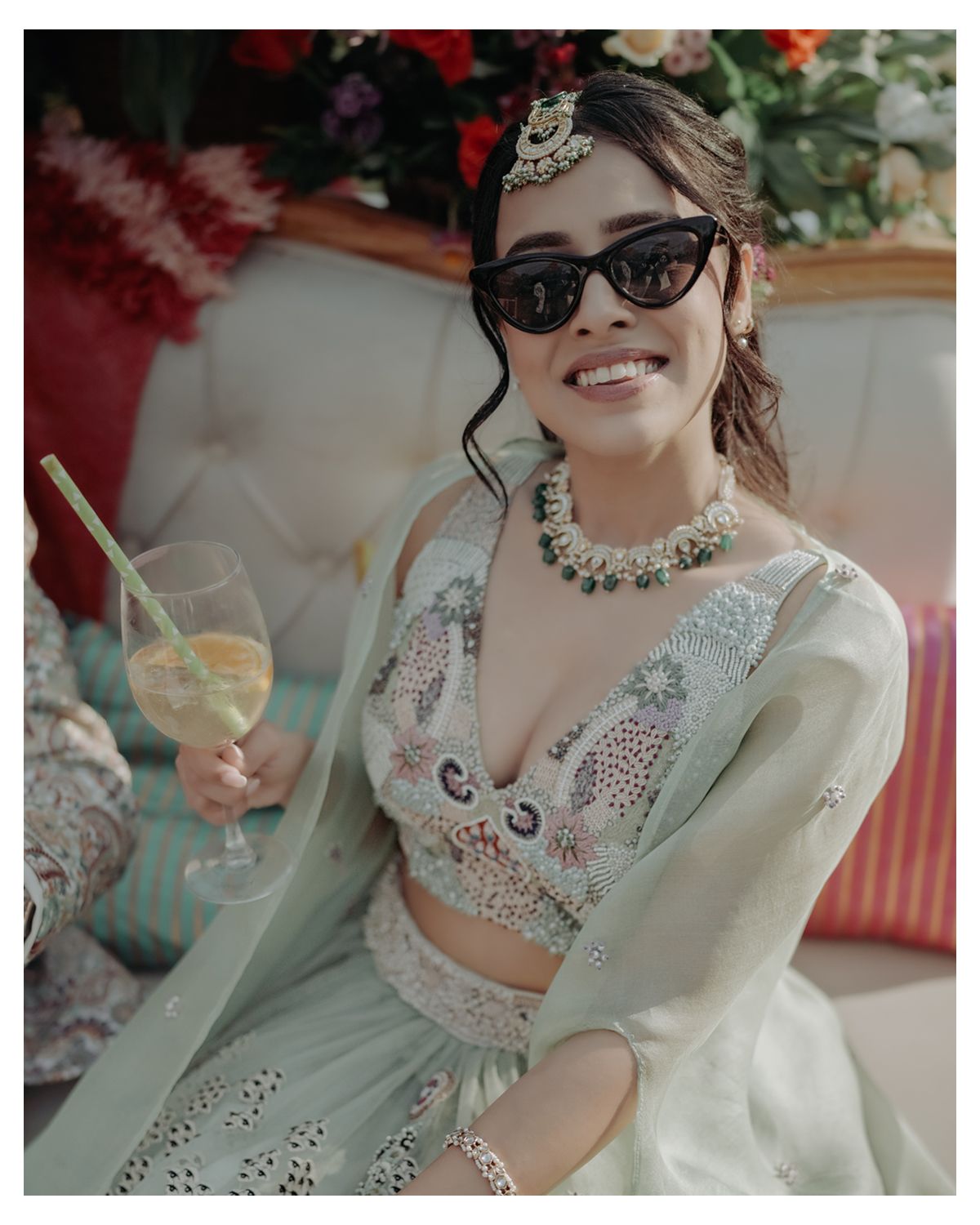 Elegant bride in mint green lehenga and sunglasses holds cocktail while relaxing at reception celebration.