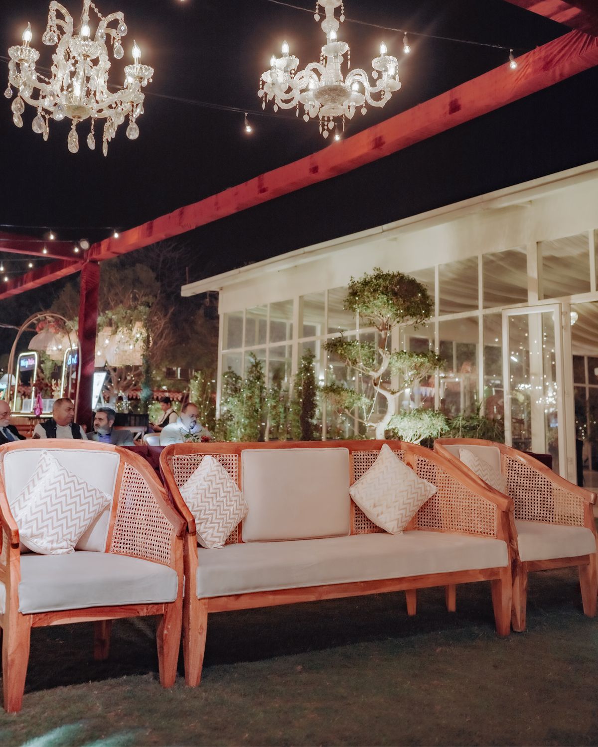 Elegant outdoor lounge area with crystal chandeliers, rattan seating, and white cushions against a glass-walled backdrop.