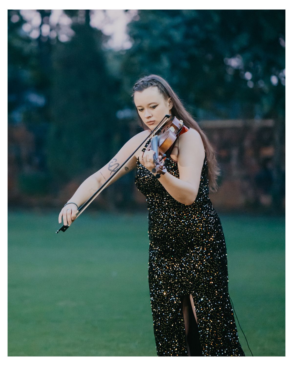 A violinist in a glittering black dress performs outdoors on a green lawn at dusk.