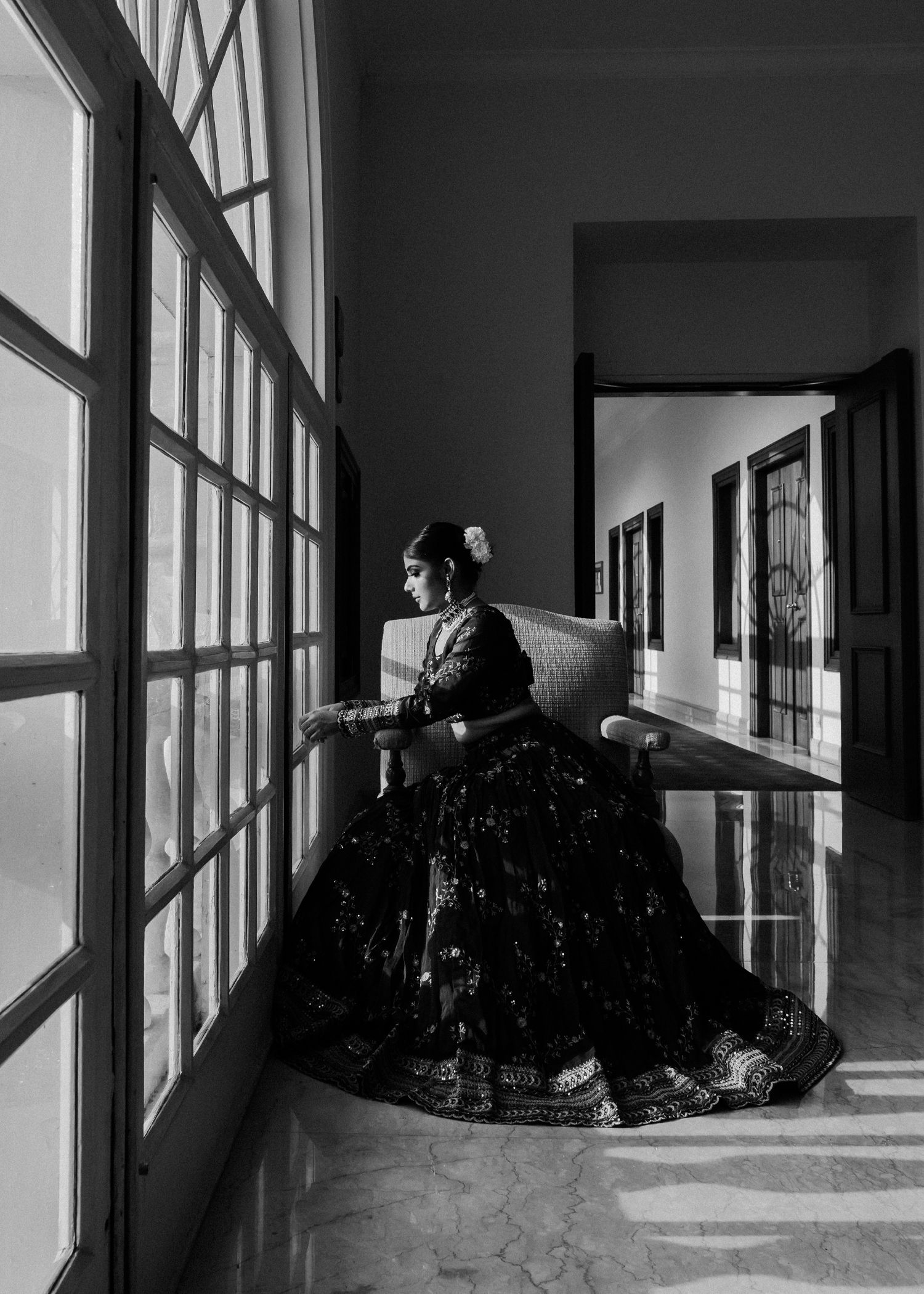 A silhouetted figure in an elegant full-skirted dress stands at a tall window in a black and white artistic photograph.