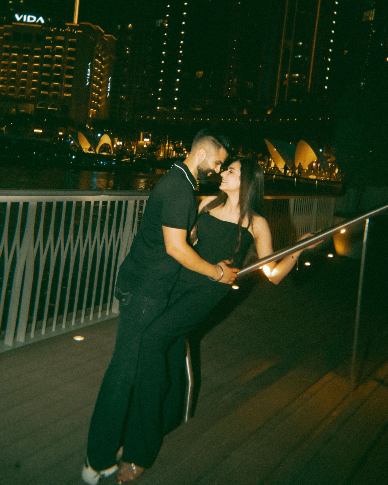 Couple kissing on a waterfront bridge at night with city skyline and Sydney Opera House in background.
