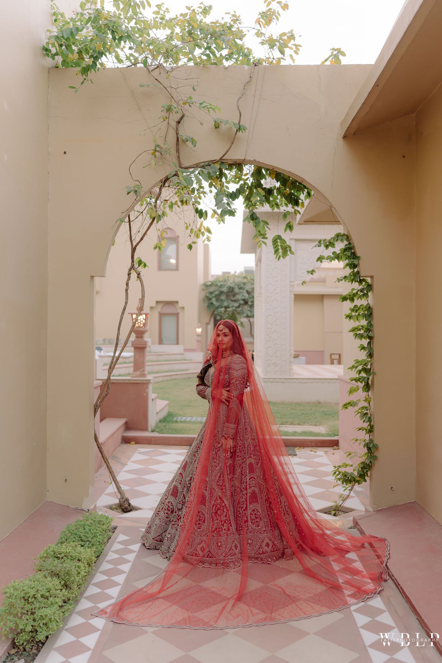 Bride in red bridal lehenga with dupatta poses under arched doorway at elegant Indian wedding venue.