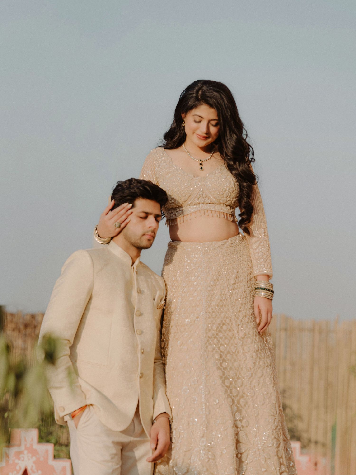 A couple in elegant gold Indian wedding attire pose outdoors, the woman standing as the man sits beside her.