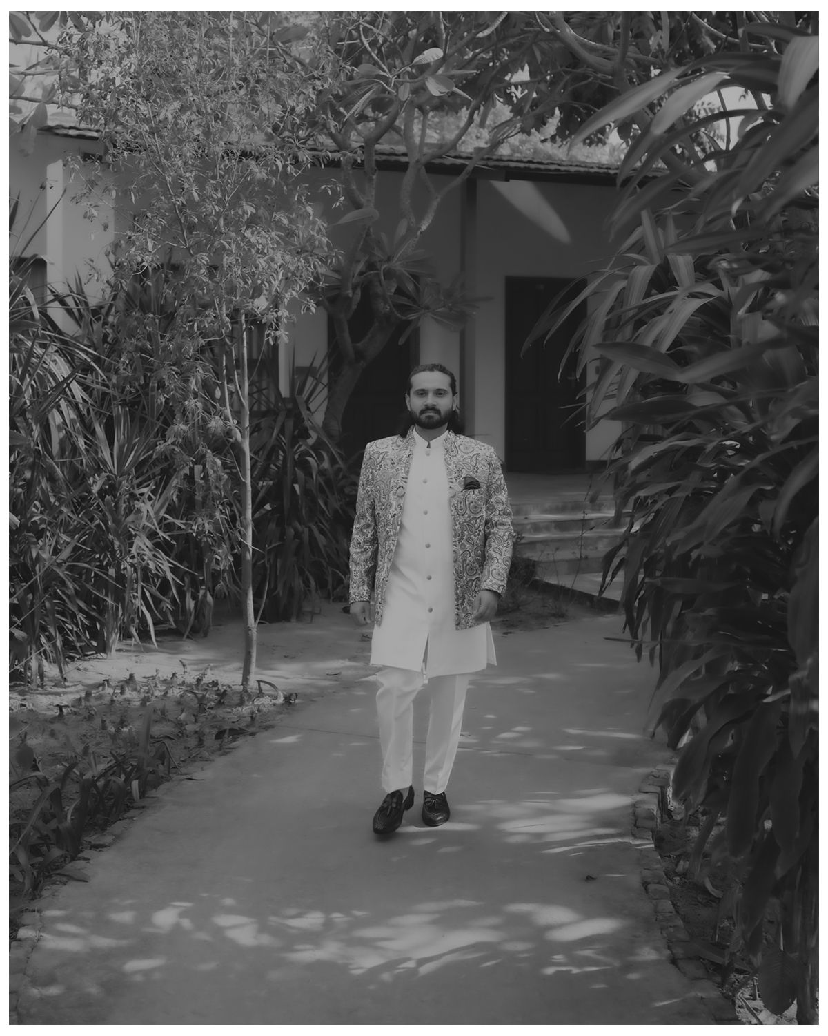 A vintage black and white photograph showing a person in light-colored attire standing on a garden path lined with plants.
