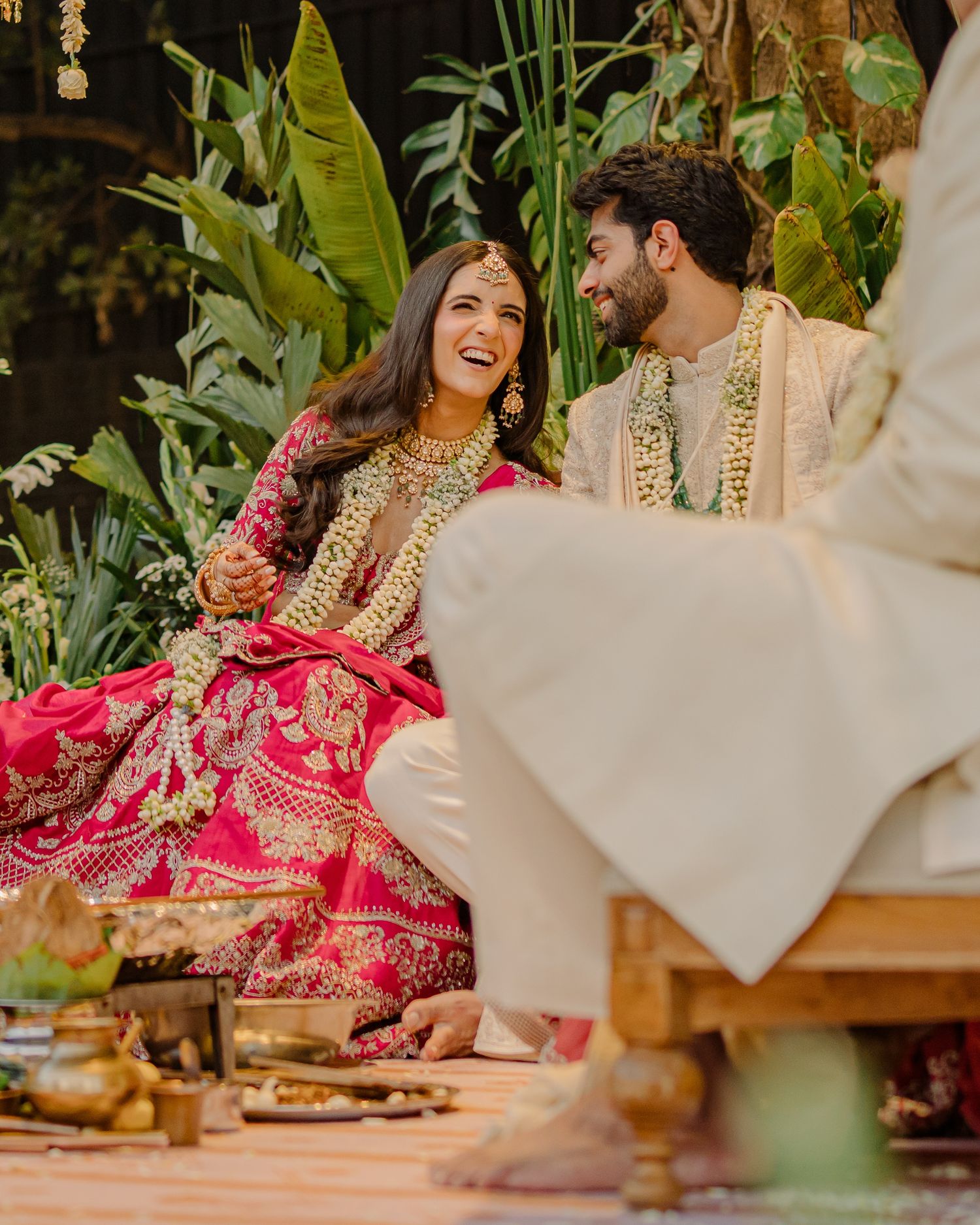 Indian bride in red lehenga and groom in white sherwani laughing together during traditional wedding ceremony.