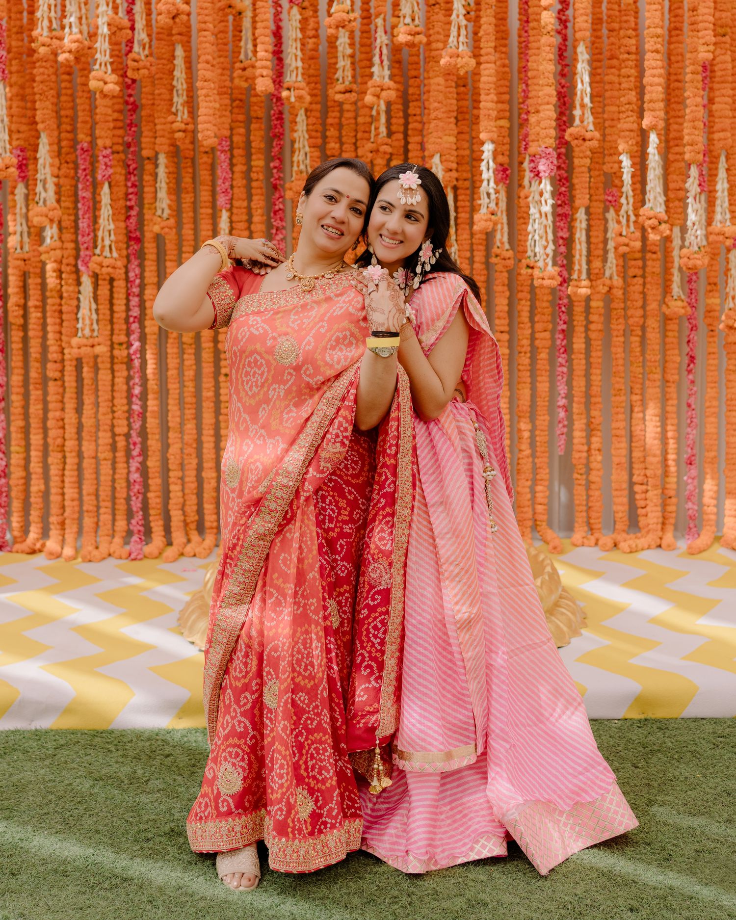 Two women in pink Indian lehengas pose together at a festive event with marigold flower backdrop.