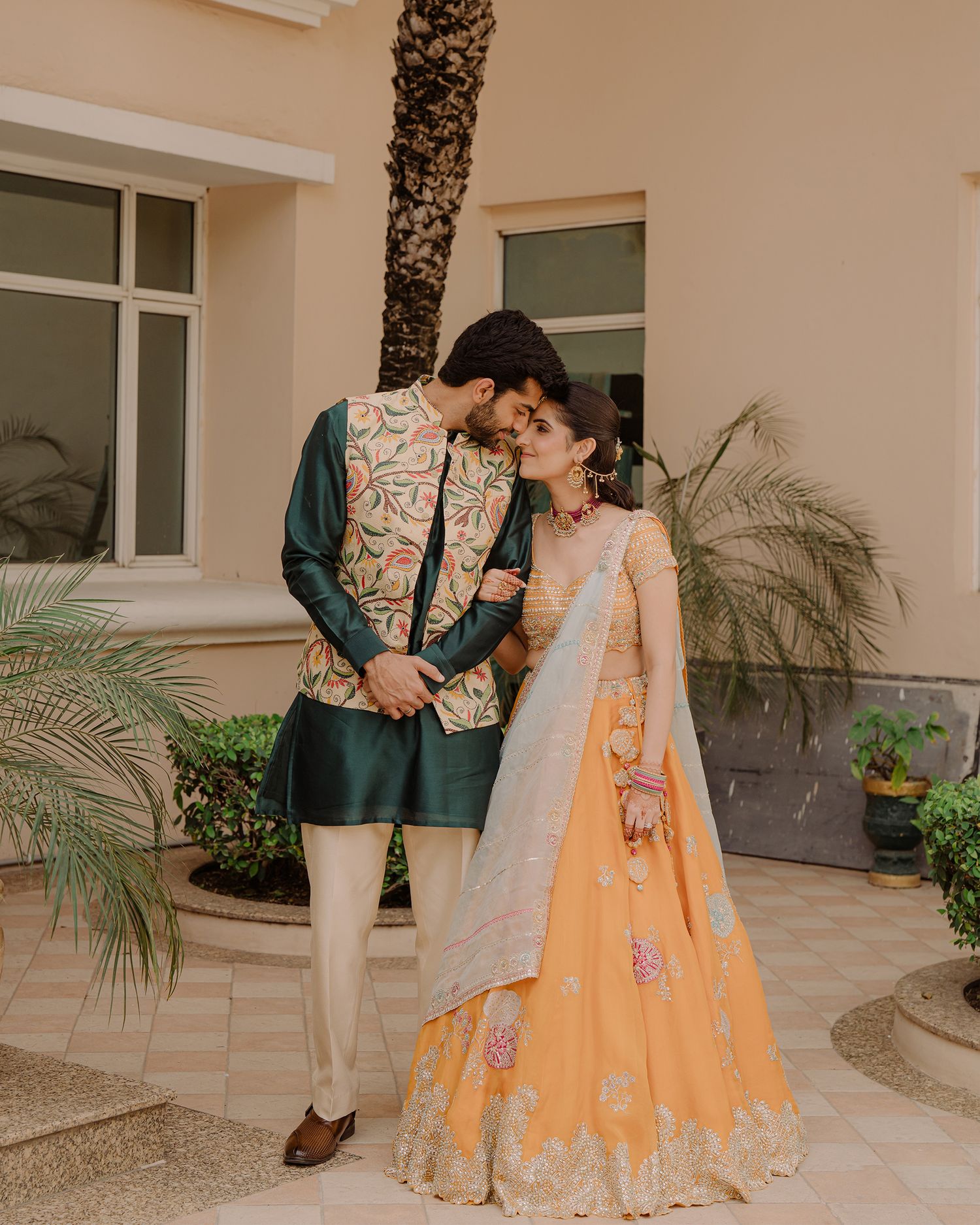 Couple in traditional Indian attire, man in green sherwani, woman in peach lehenga, posing outdoors.