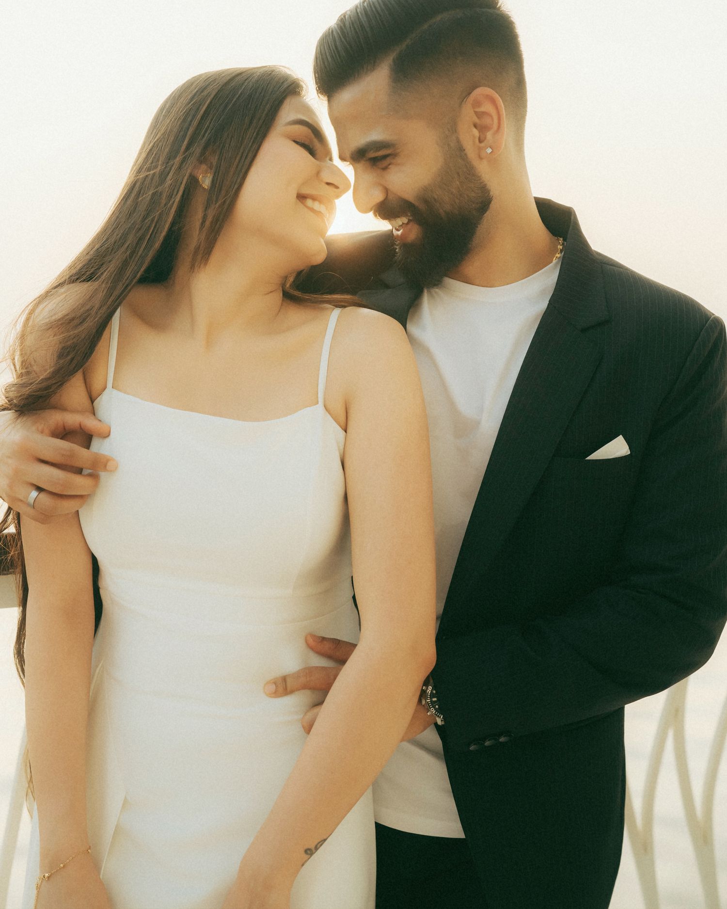 A couple in white dress and black suit smiling at each other in a romantic outdoor pre-wedding photoshoot.