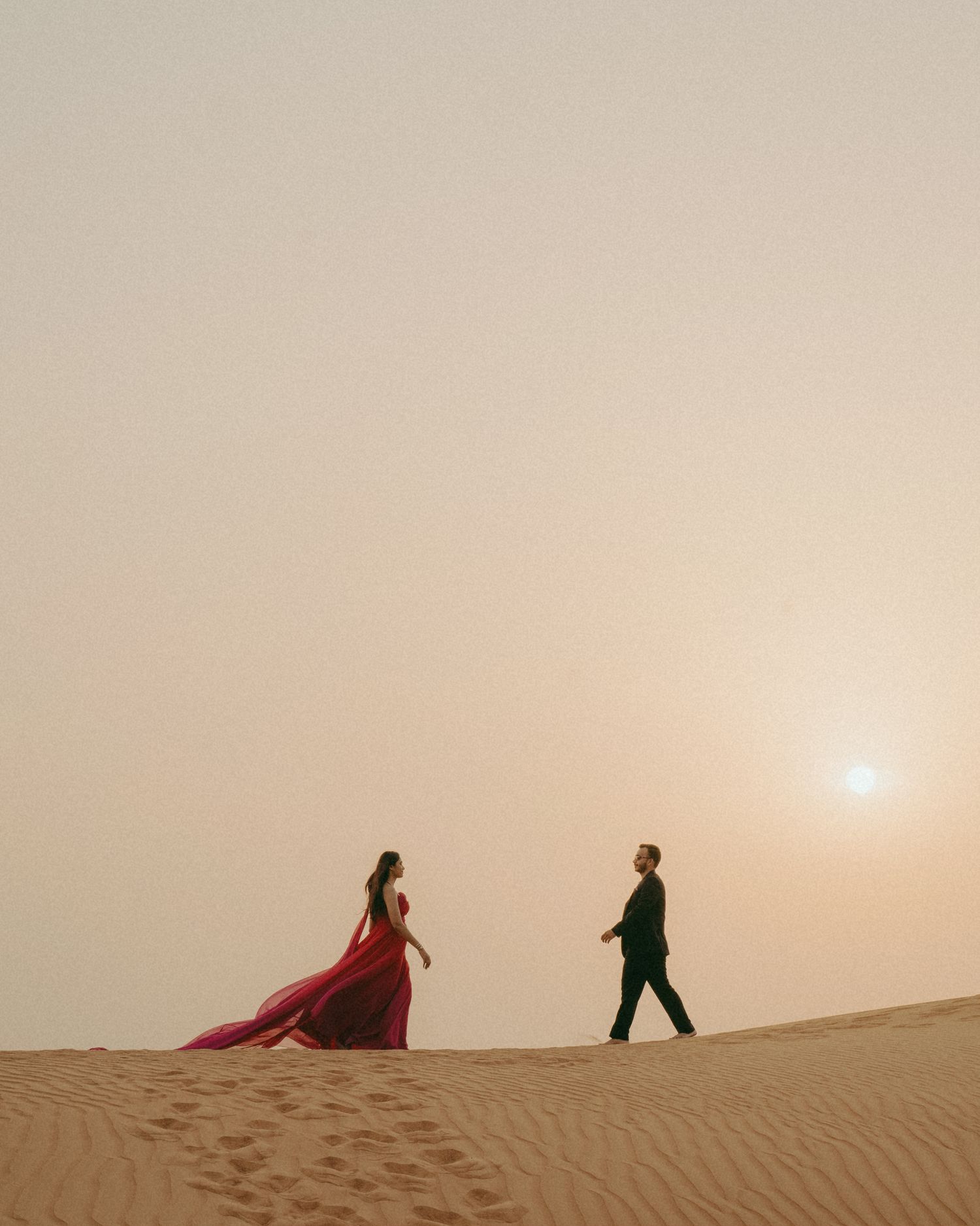 Couple in red gown and suit walking on desert sand dunes at sunset in a romantic editorial photo.