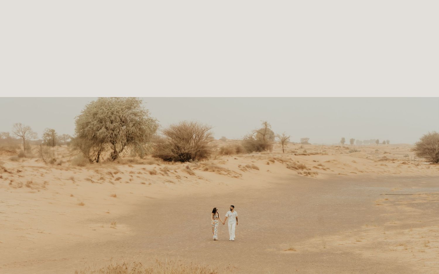 Two people in white attire walk hand in hand through a vast sandy desert landscape with sparse trees.