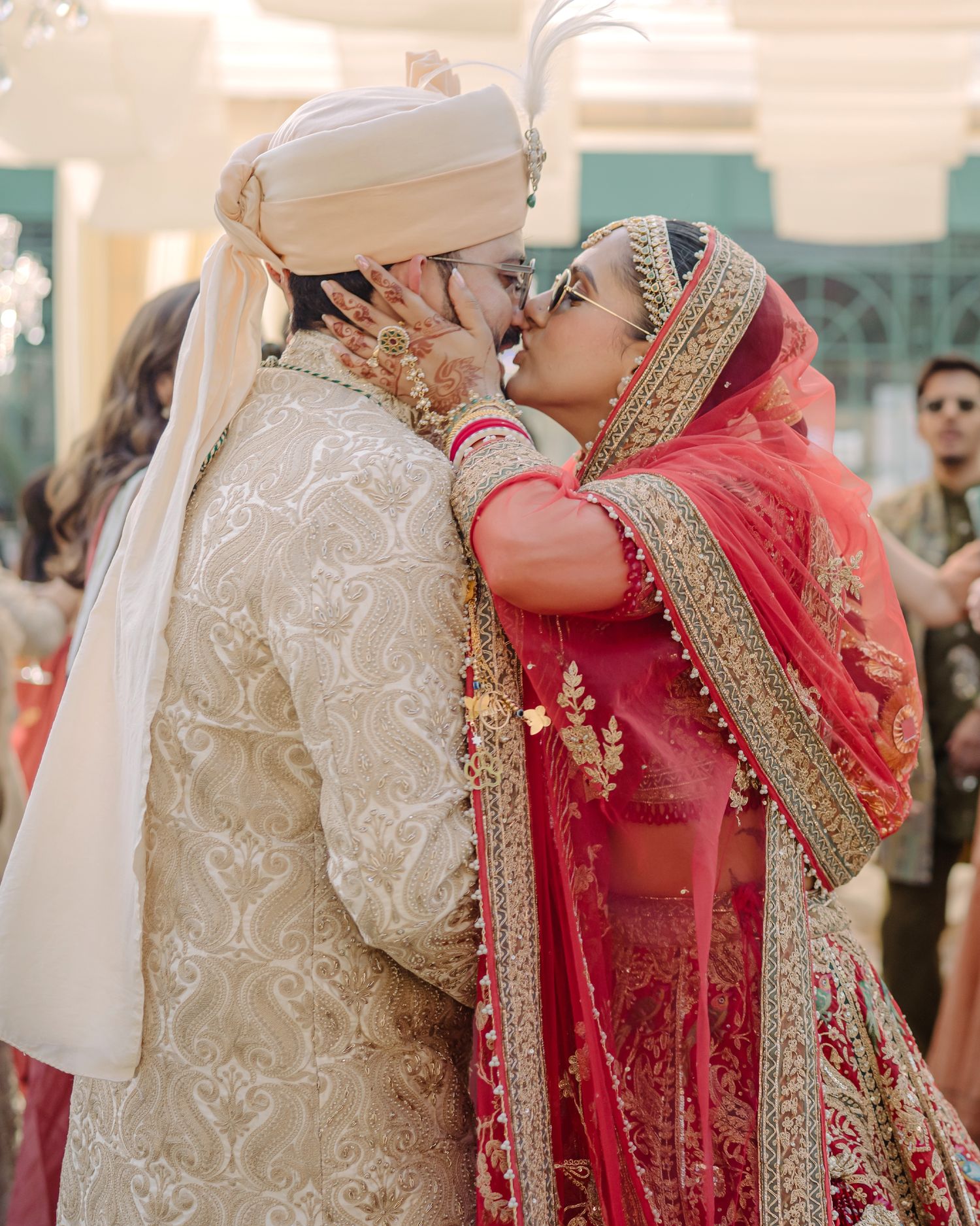 Indian bride and groom share a kiss during traditional wedding ceremony, wearing ornate red and gold bridal attire.