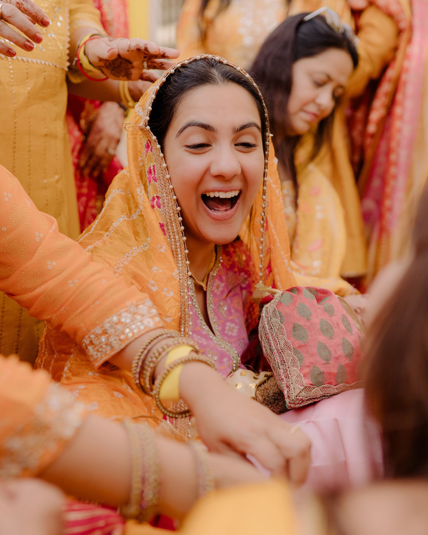 A joyful bride in a pink and orange lehenga laughs during a traditional Indian wedding ceremony surrounded by guests.