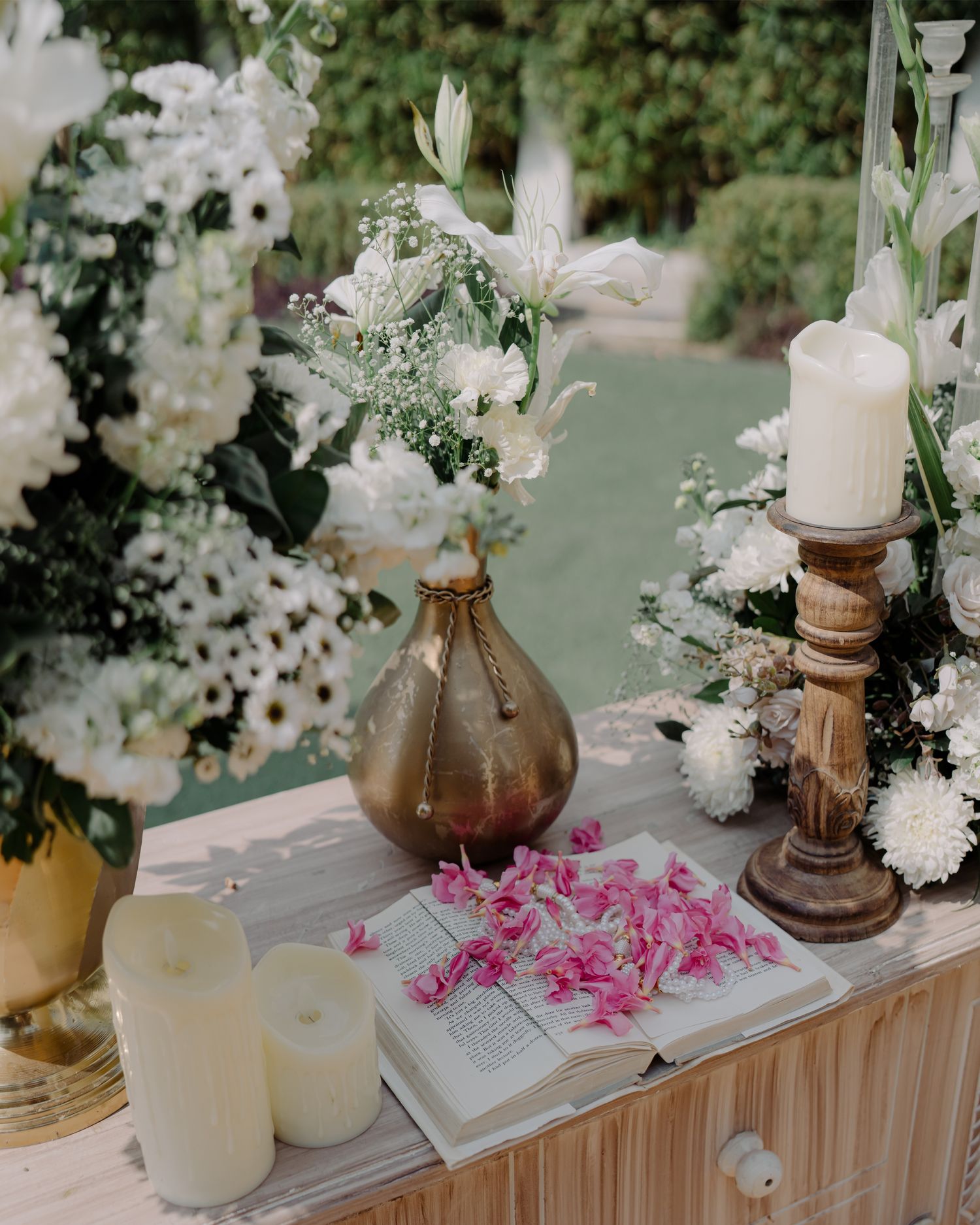 Elegant wedding table with white floral arrangements, candles, open book, pink petals, and a brass vase on a wooden surface.