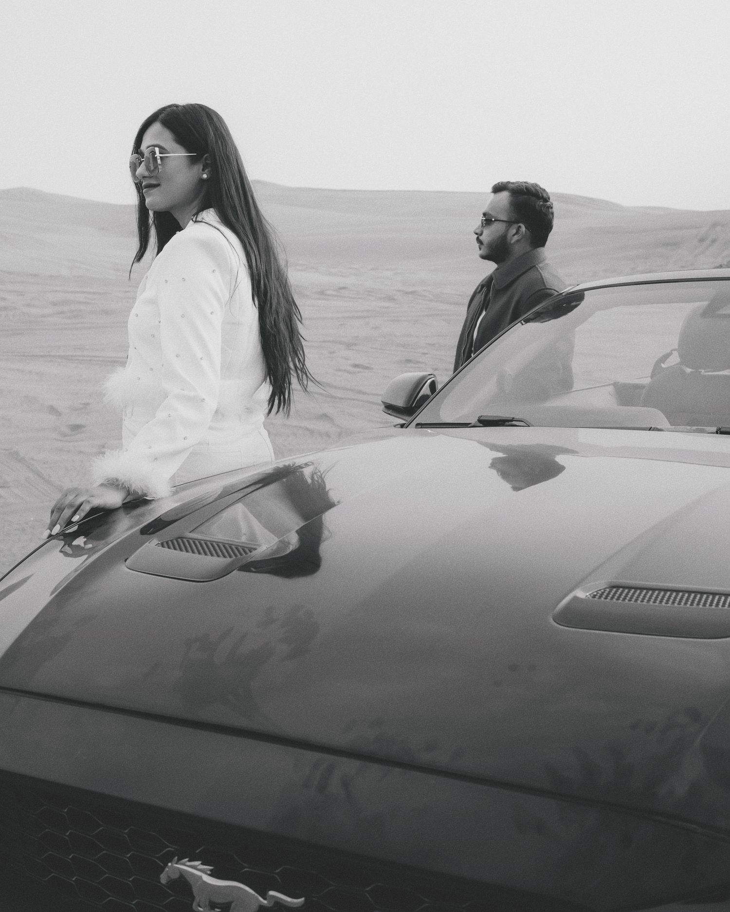 Black and white photo of a couple posing by a classic car in a desert landscape.