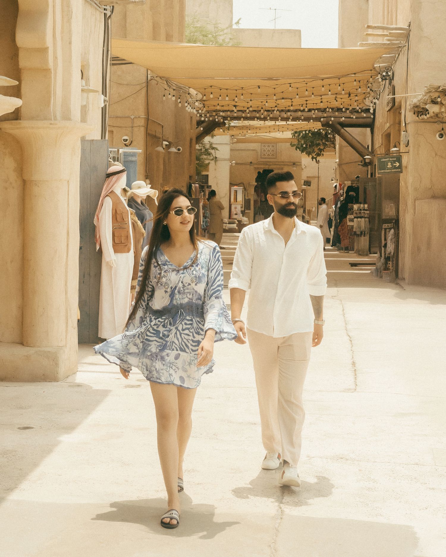 Couple walking hand in hand through a sunny Middle Eastern marketplace with stone architecture and string lights.