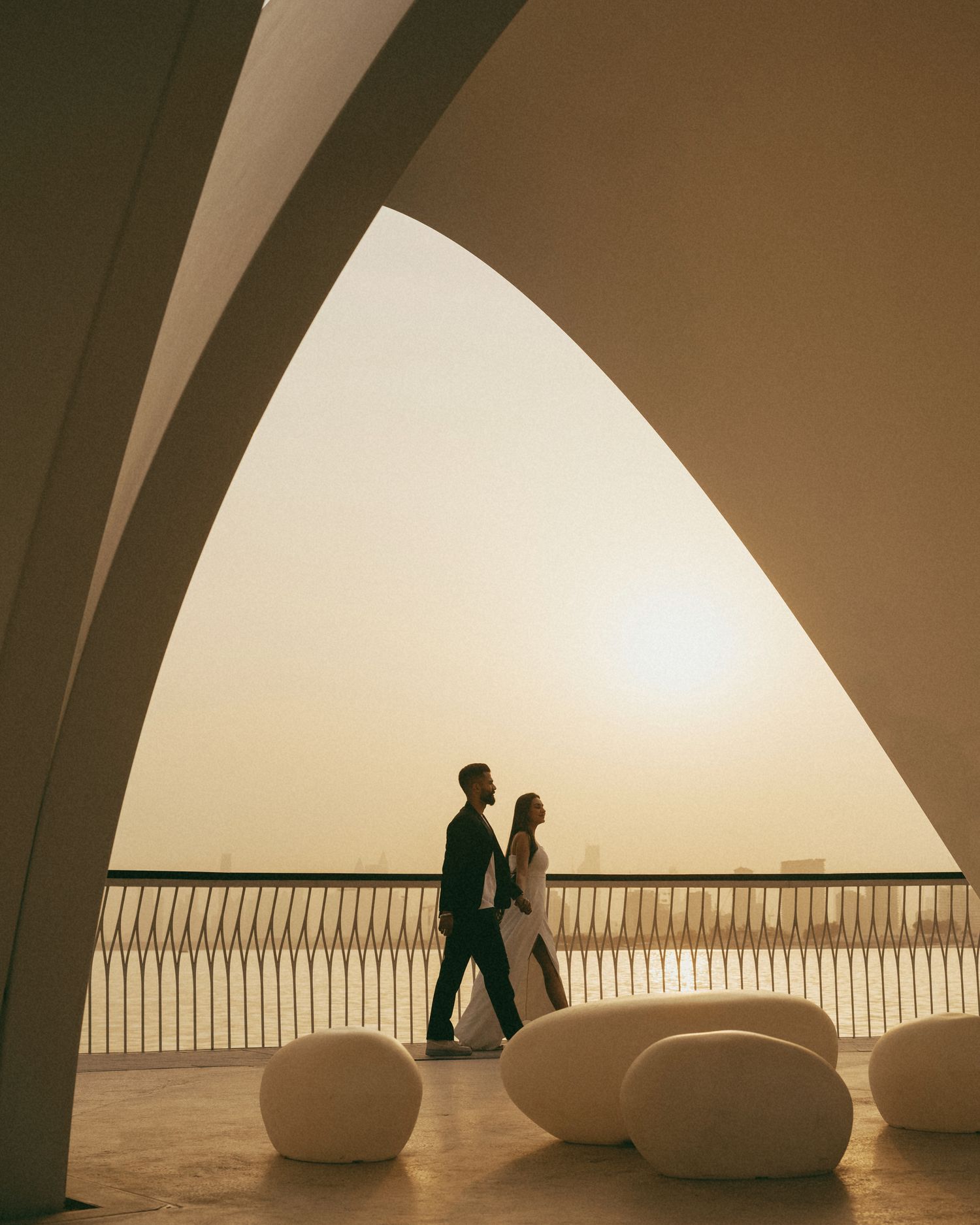 Couple silhouetted against golden sunset under elegant white arches on a modern terrace with pebble-shaped seats.