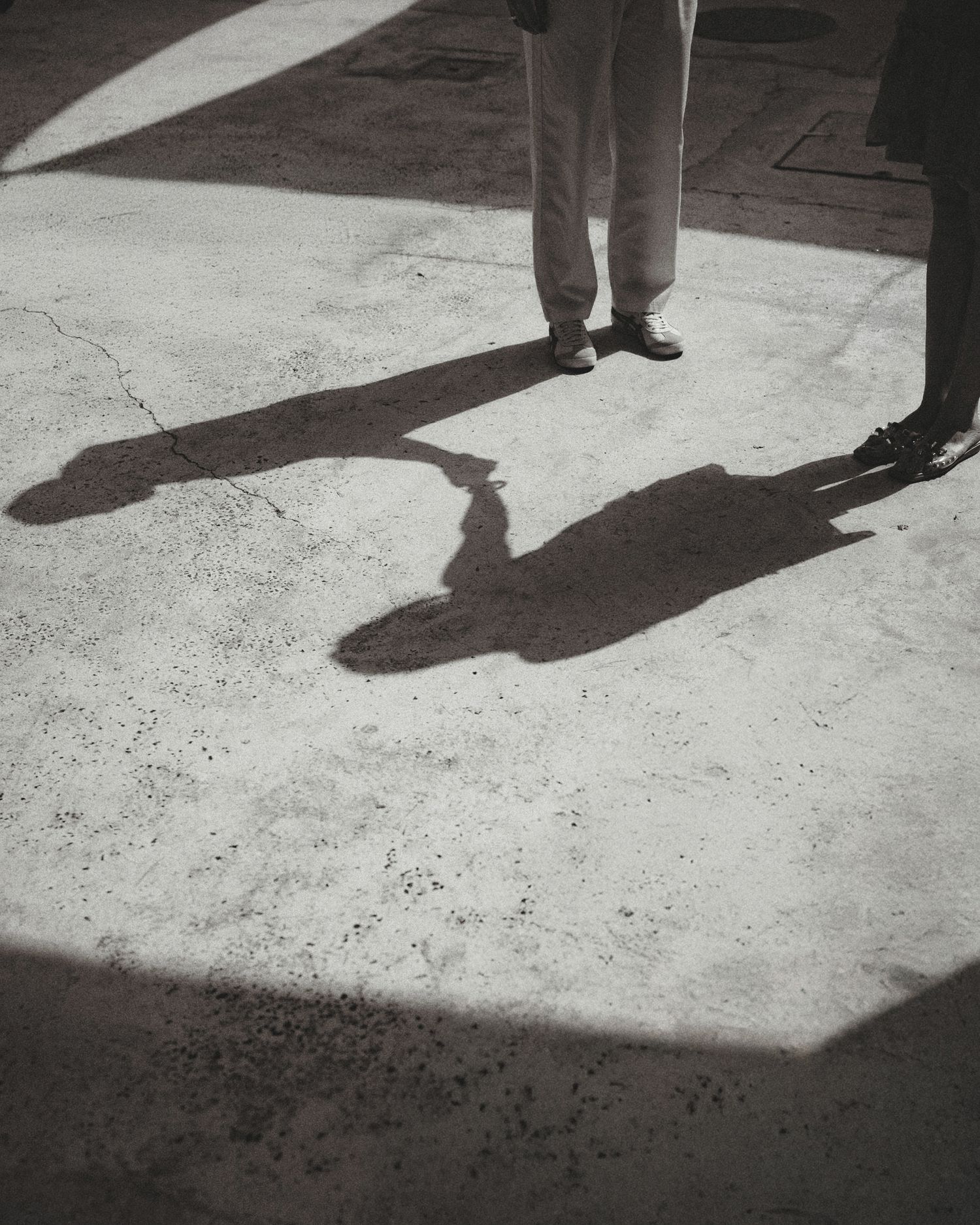 Black and white photo of two people standing on concrete, their long dramatic shadows stretching across the sunlit ground.
