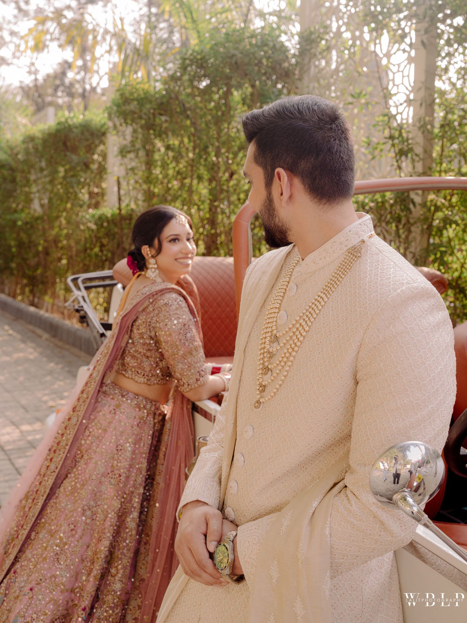 Indian bride in embroidered lehenga and groom in cream sherwani pose near vintage car at outdoor wedding.
