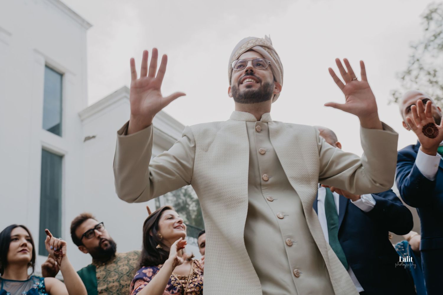 Groom in cream sherwani raises hands joyfully during outdoor wedding celebration.